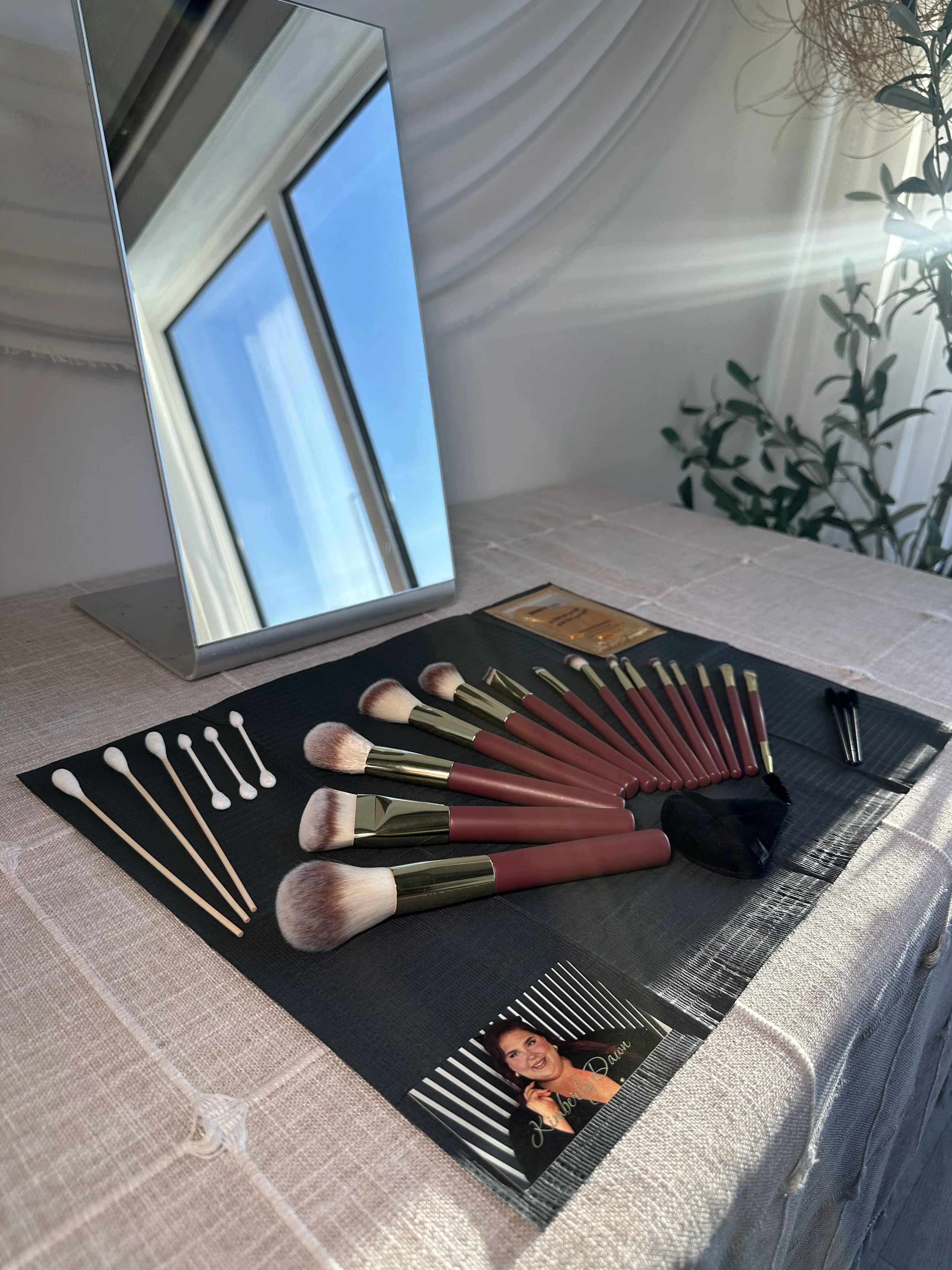 A set of makeup brushes and tools arranged on a black mat, with a mirror and a picture of a woman in the background, on a white tablecloth.