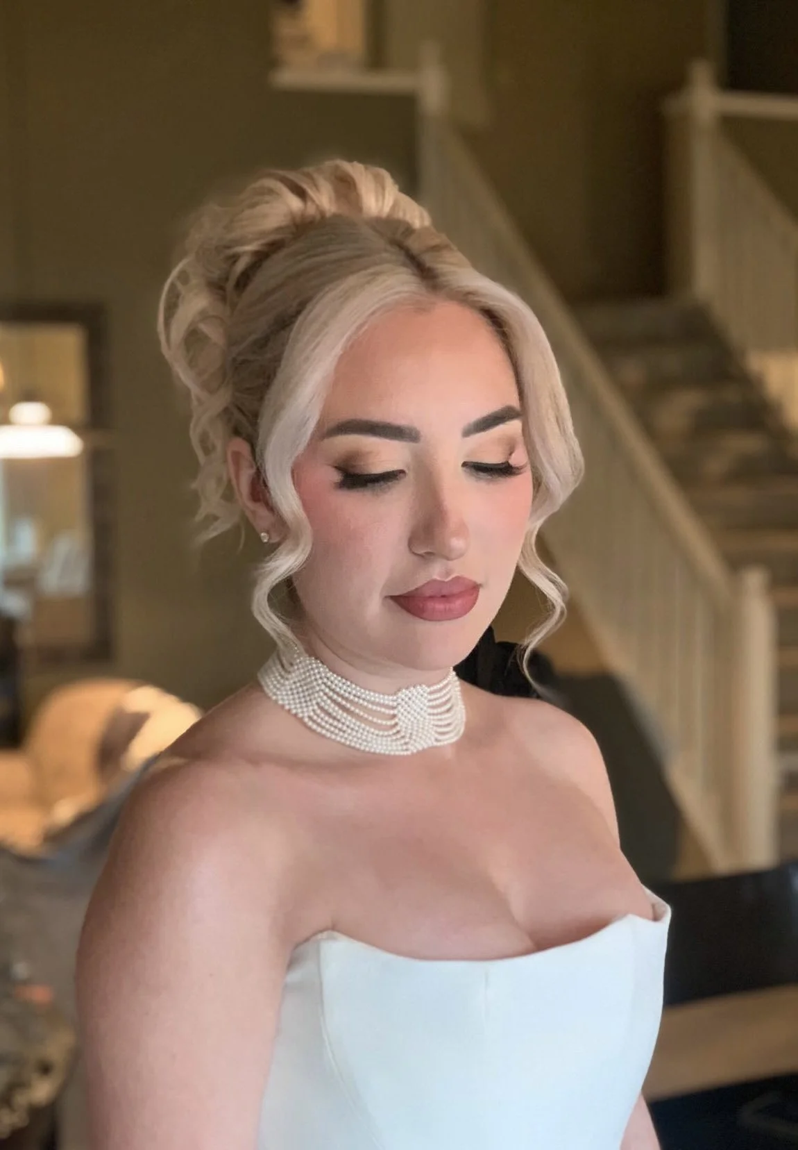 A woman with platinum blonde hair styled in an updo with loose curls, wearing a pearl choker necklace and a strapless white dress, with makeup featuring nude shades and a soft smile, standing indoors with a staircase in the background.