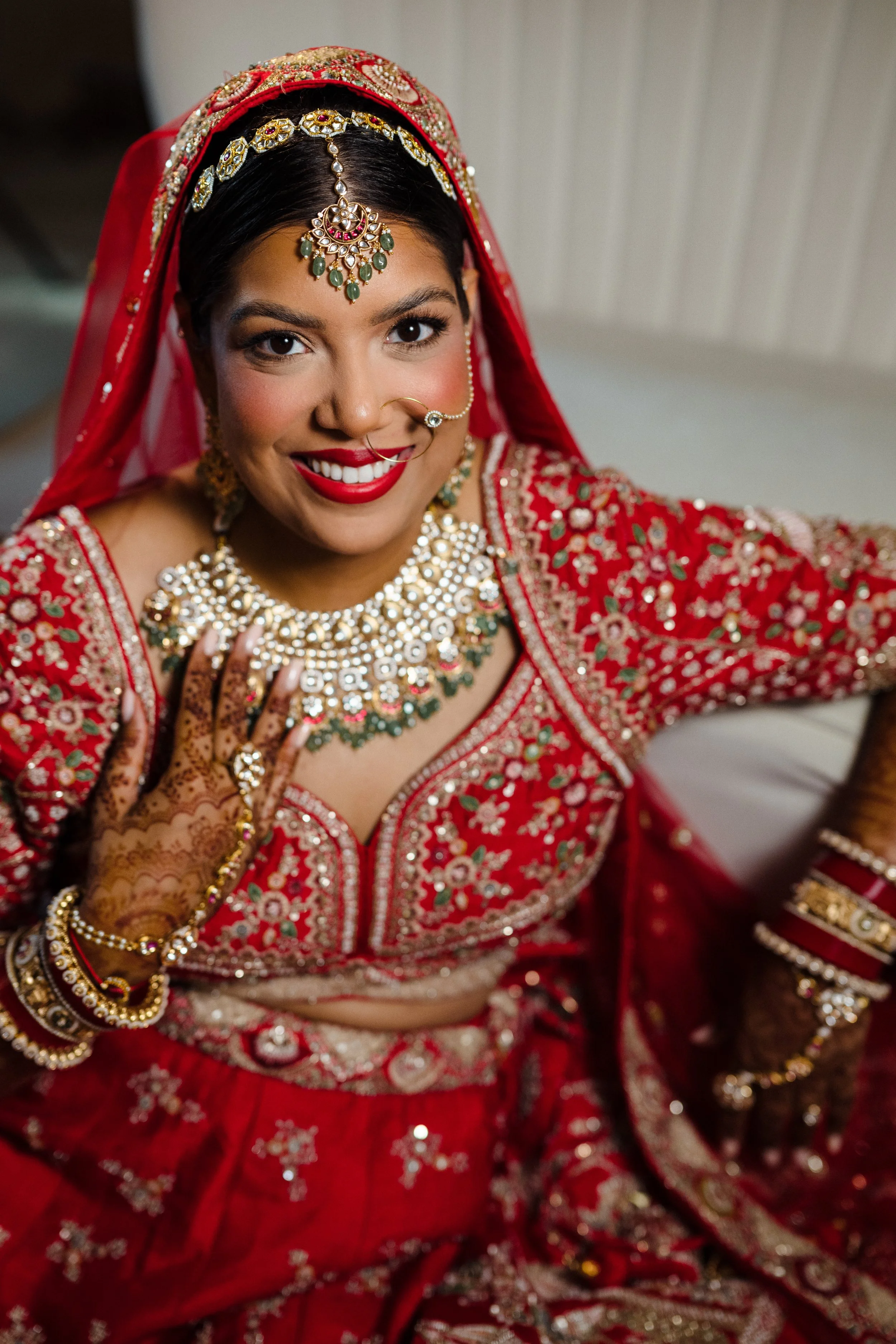 A woman dressed in elaborate red traditional Indian bridal attire with gold, green, and pearl jewelry, including a maang tikka, nose ring, necklace, earrings, and bangles, smiling at the camera.