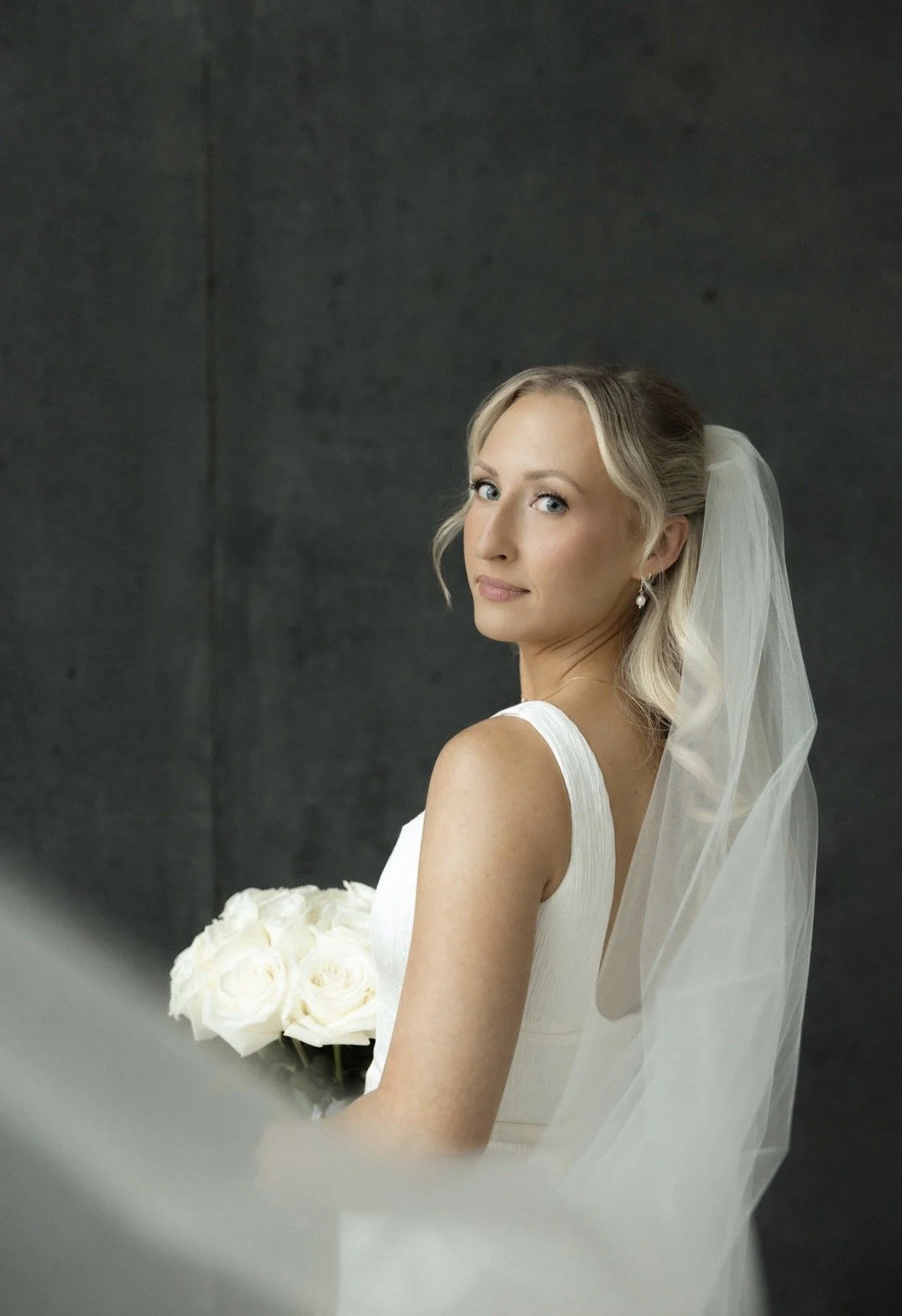 A bride with blonde hair in loose waves, wearing pearl earrings, a white wedding dress, and a veil, is holding a bouquet of white roses and looking over her shoulder at the camera against a dark gray background.