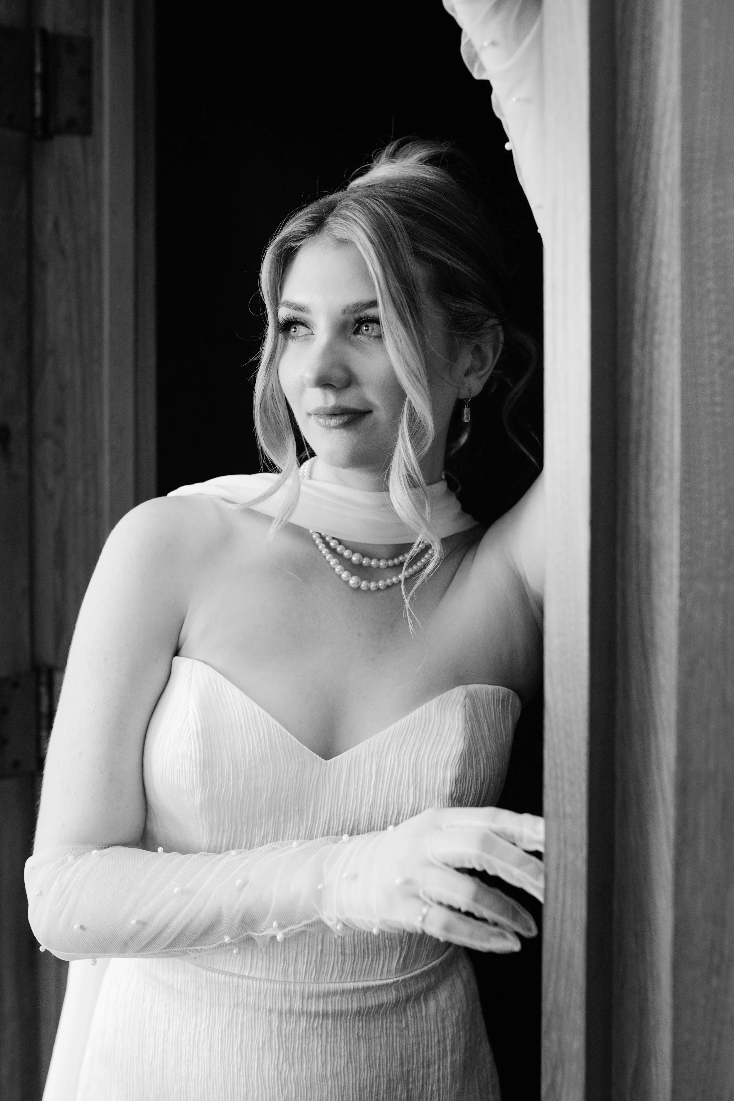 A black and white photograph of a woman in a strapless dress with pearl jewelry, standing near a doorway, looking off to the side with a serene expression.