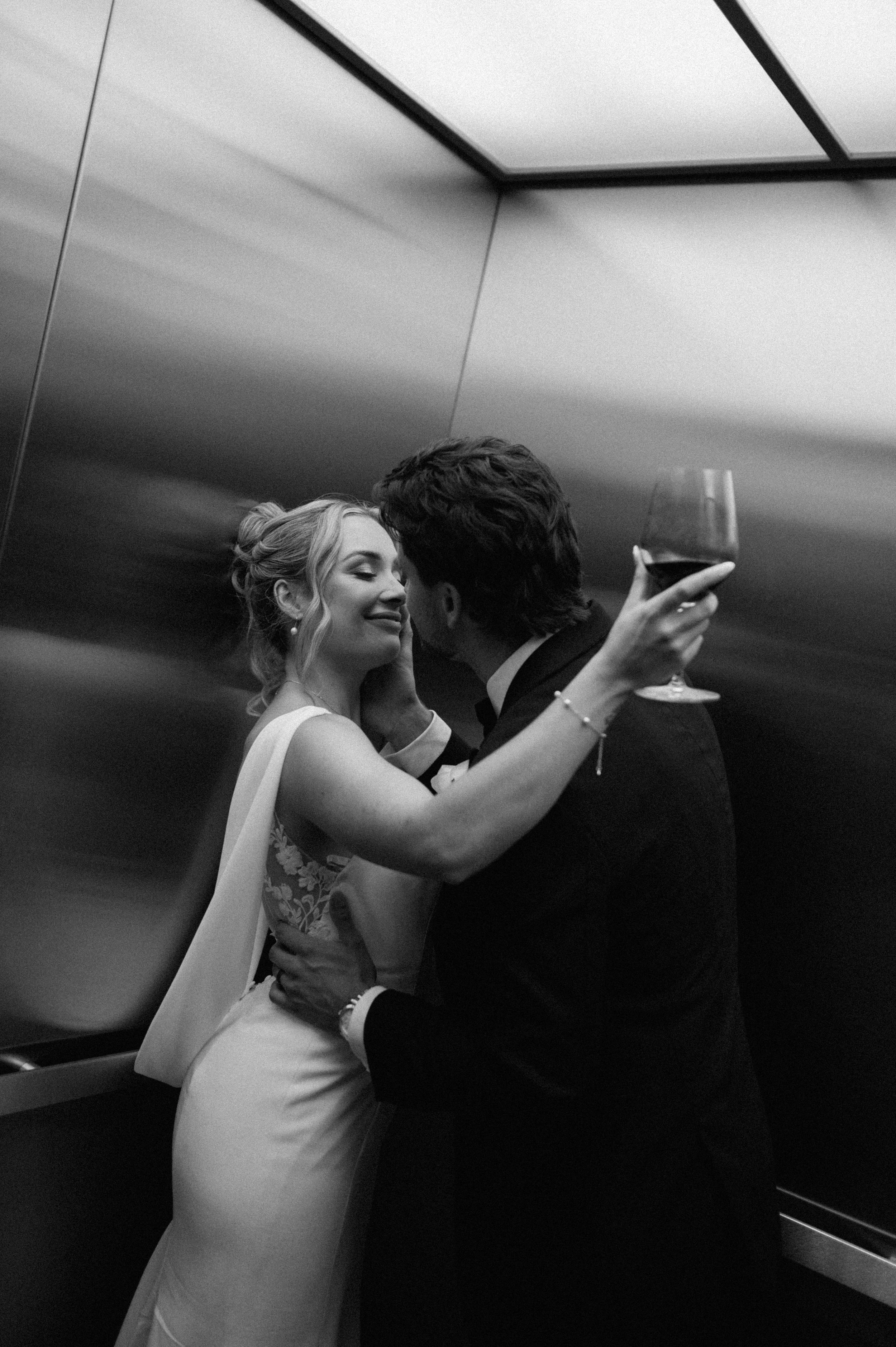 A black and white photo of a bride and groom in an elevator, sharing an intimate moment. The bride is smiling with her eyes closed, and the groom holds a glass of wine.