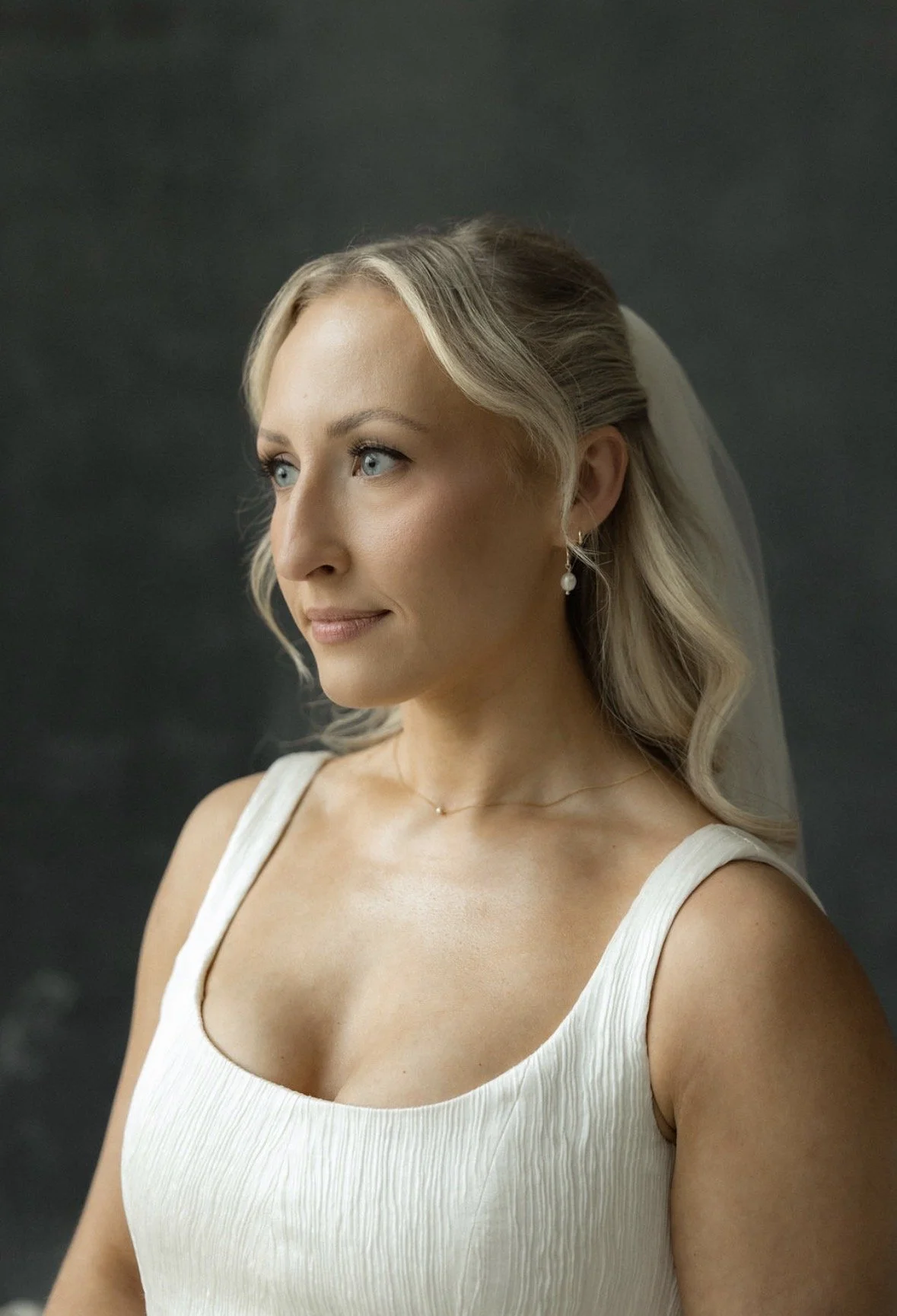 A blonde woman with blue eyes wearing a white dress, pearl earrings, and a small necklace, looking to her left with a neutral expression.