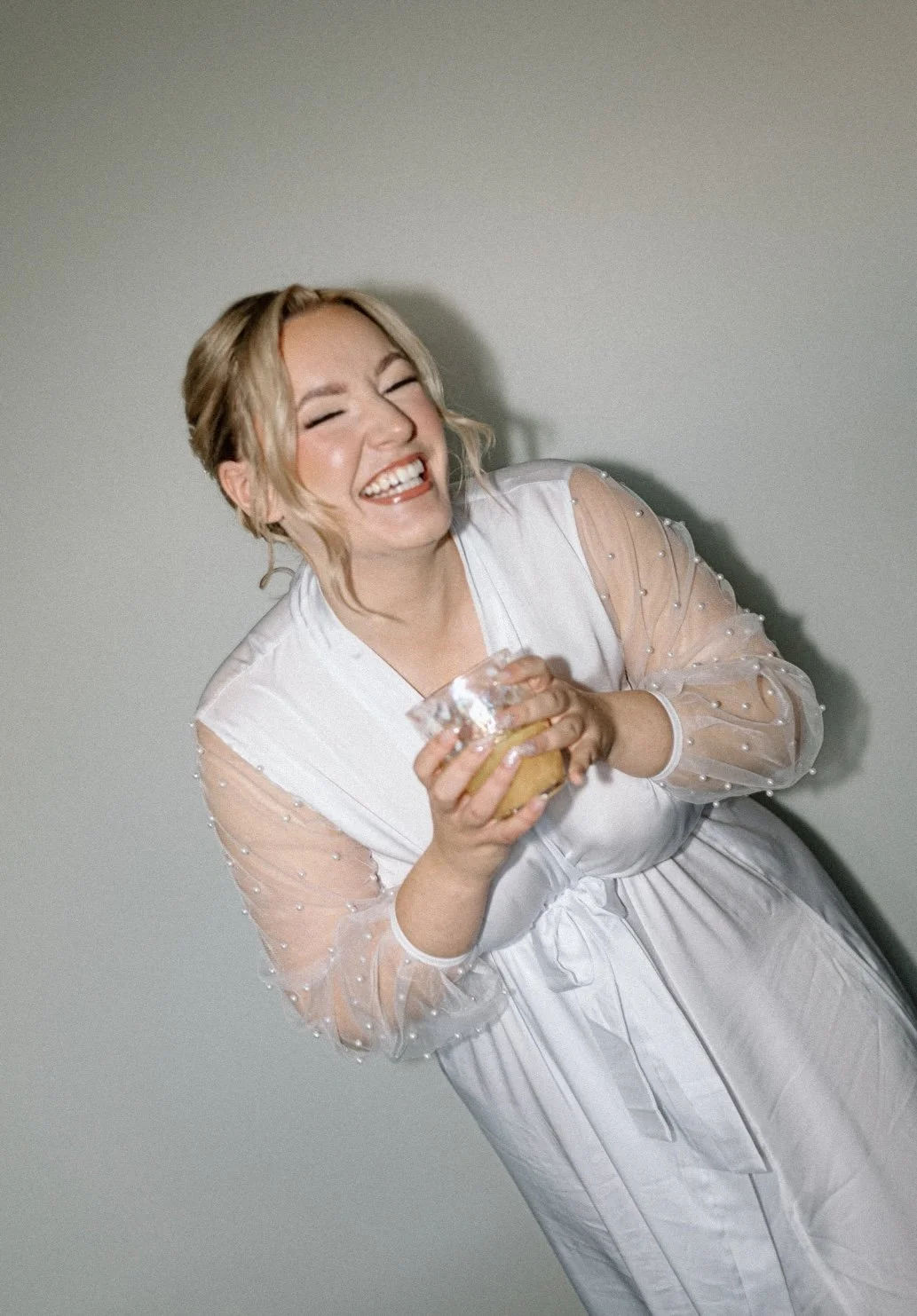 A woman with blonde hair laughing joyfully, holding a glass bowl with a snack, wearing a light-colored dress with sheer sleeves decorated with small pearls.