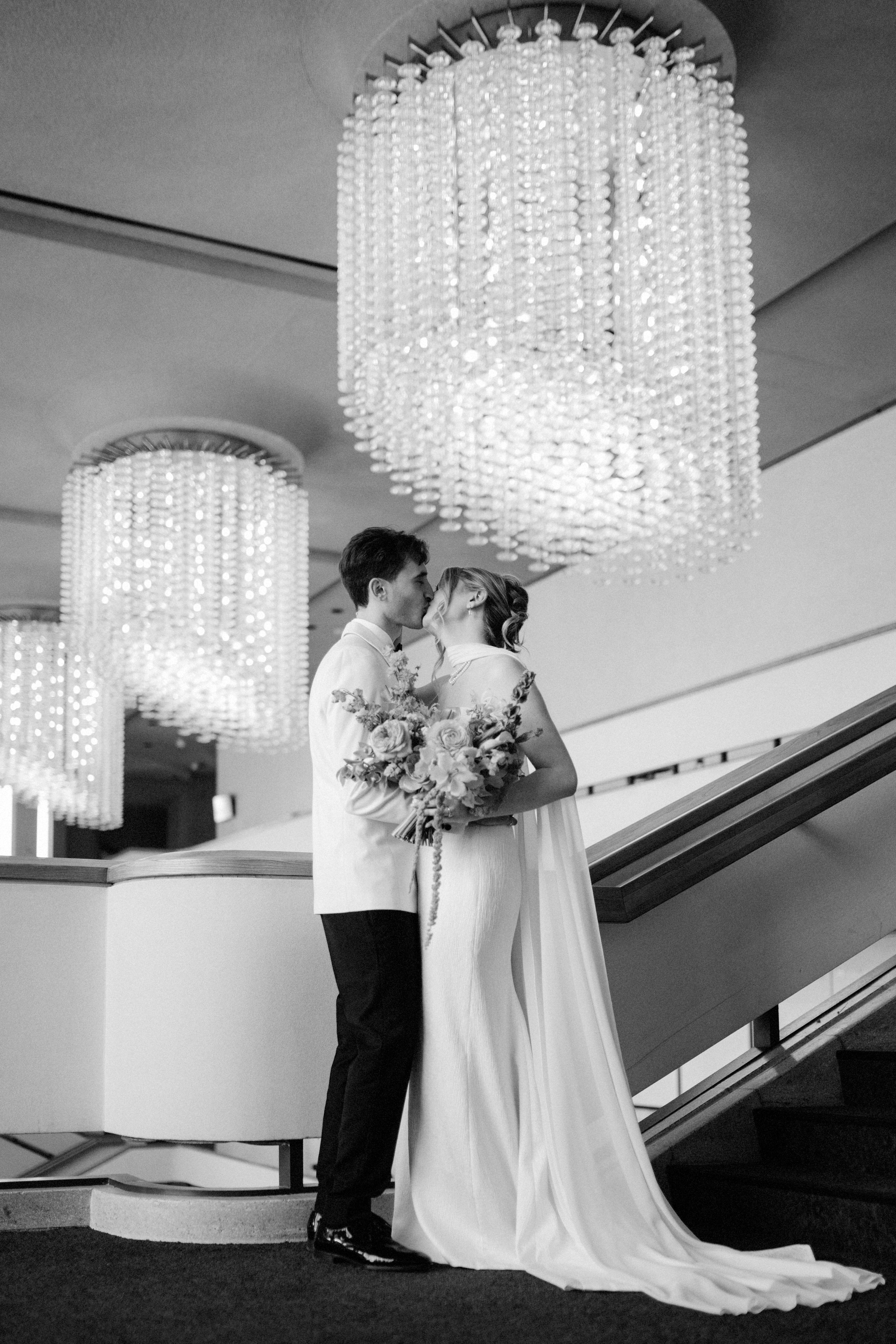 A black and white photograph of a bride and groom sharing a kiss on a staircase. The bride is holding a large bouquet of flowers and wearing a long, flowing gown. The groom is dressed in a suit. Large ornate chandeliers hang from the ceiling above them.