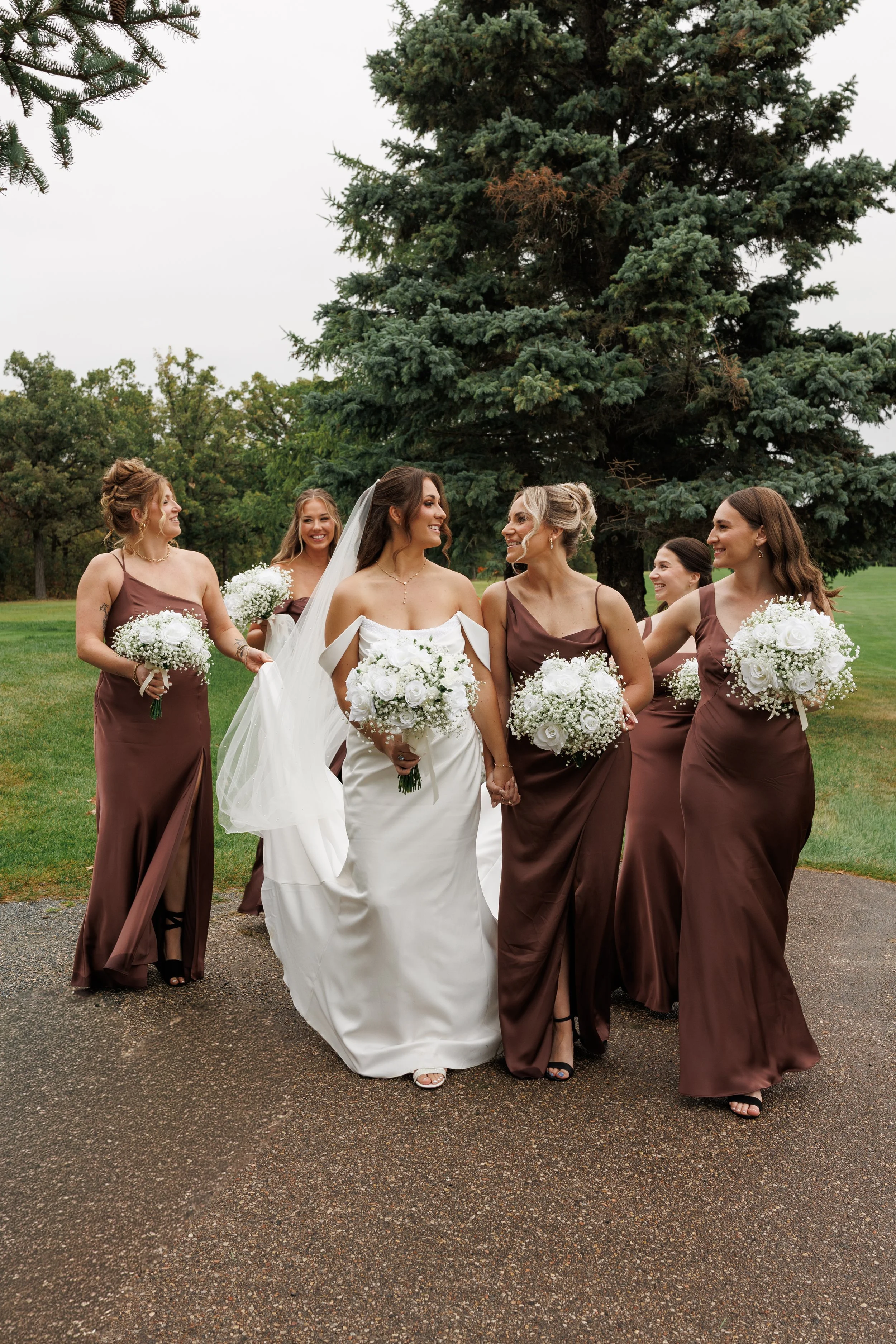 A bride in a white wedding gown holding hands with five bridesmaids in brown satin dresses, walking outdoors on a gravel path, surrounded by green grass and large trees.