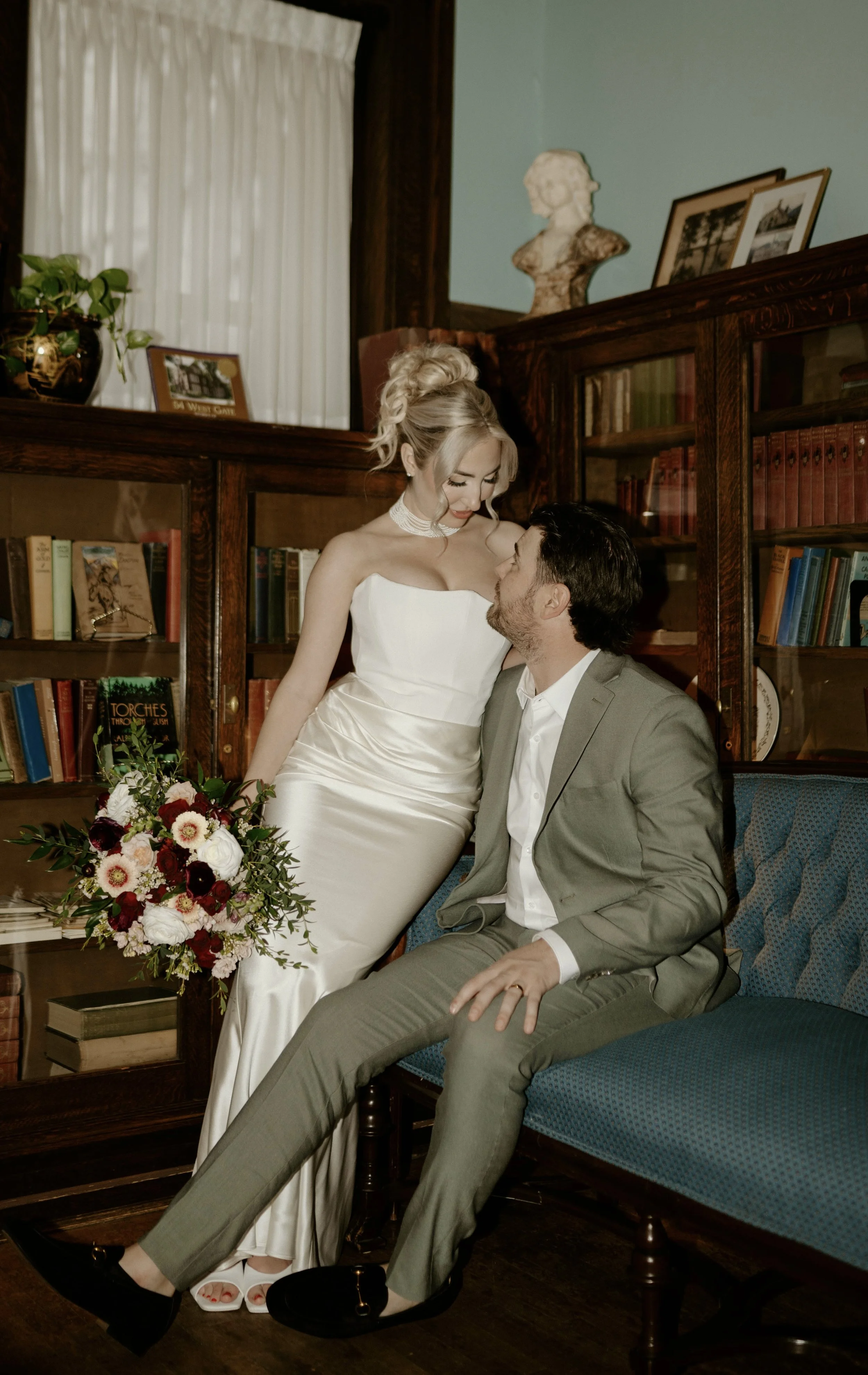 A bride in a white wedding dress standing close to a groom in a gray suit, seated on a blue upholstered sofa, in a room with bookshelf and decor. The bride is holding a bouquet of red, white, and pink flowers with green foliage, and they are looking 