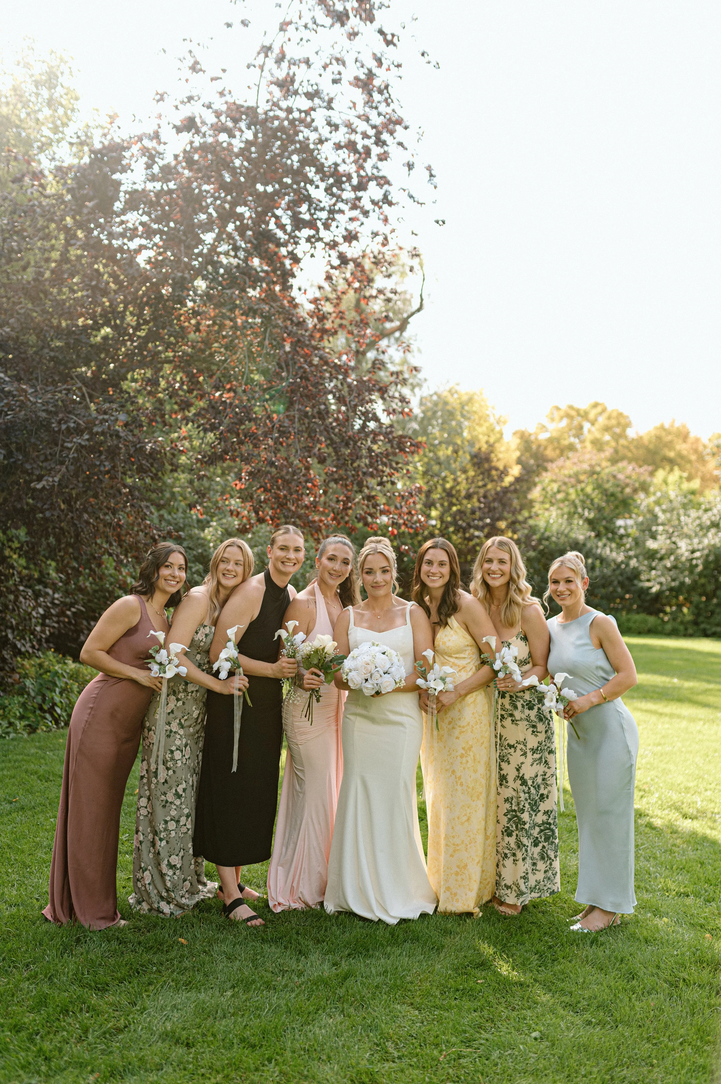 A group of nine women dressed in colorful dresses standing outdoors in a park with green grass and trees in the background, smiling at the camera, holding bouquets, with a bright sky overhead.