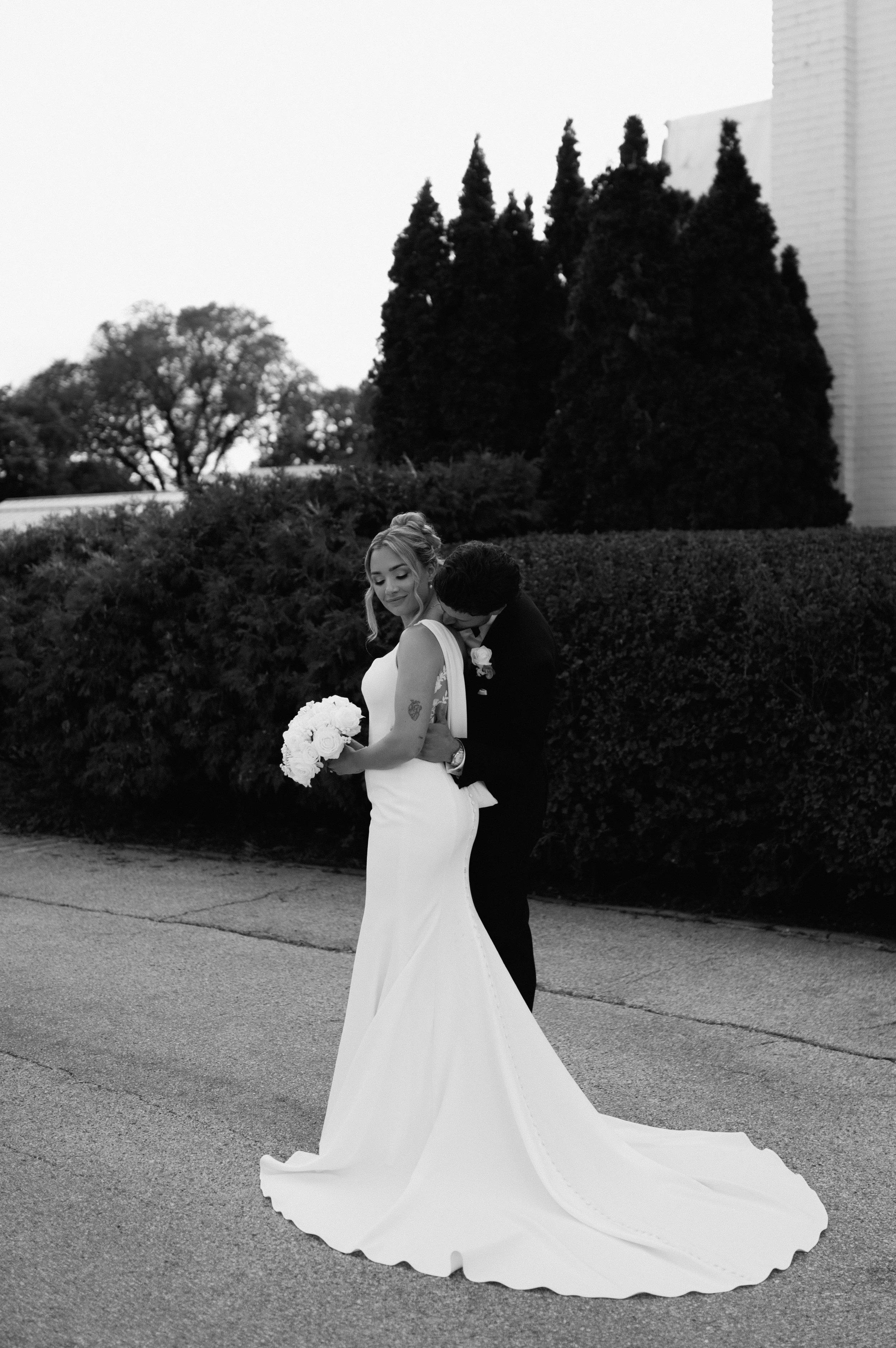 Black and white photograph of a bride in a long white gown holding a bouquet, with a groom standing behind her, outdoors with a hedgerow and tall trees in the background.