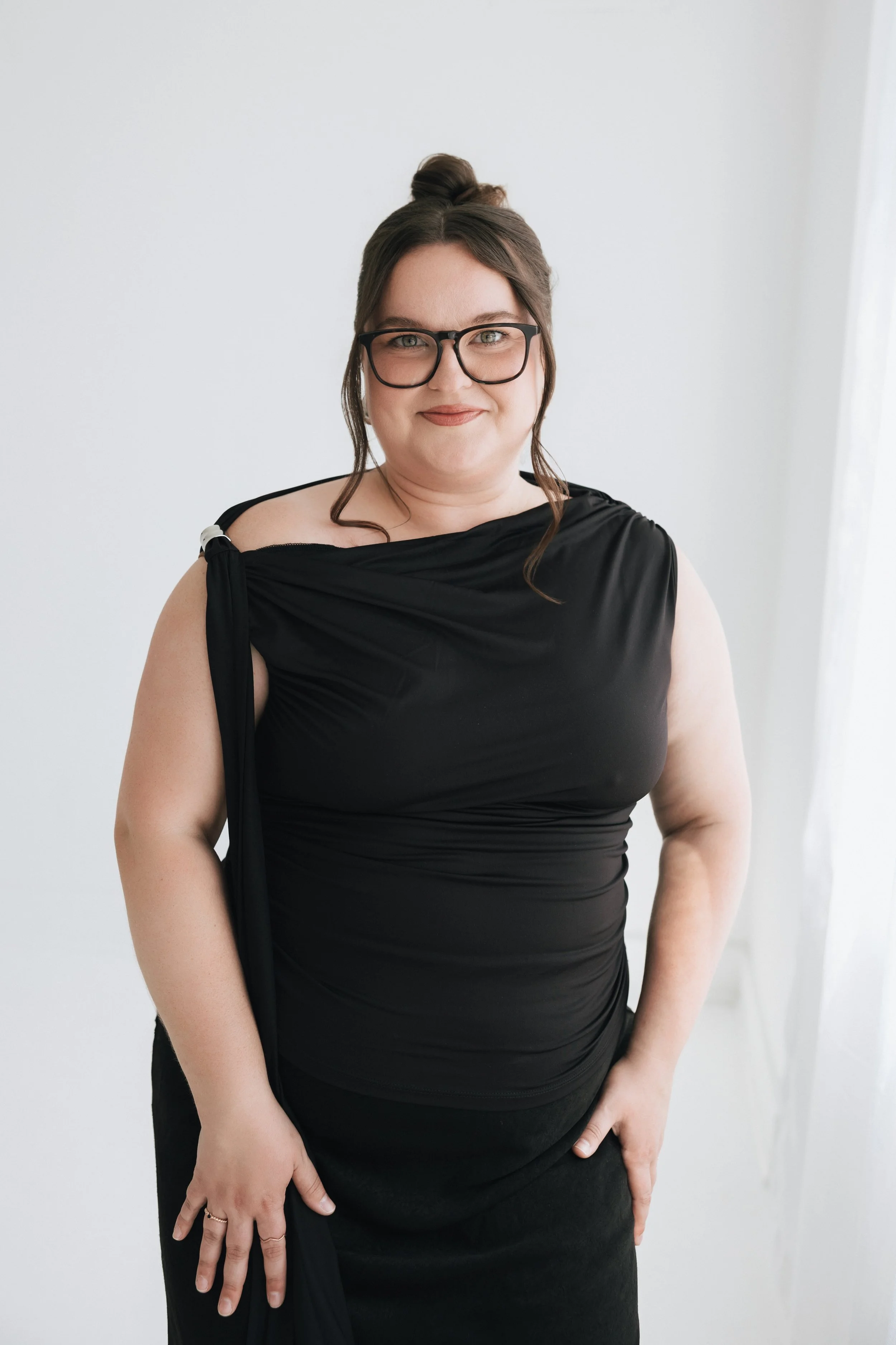 A woman with glasses wearing a black dress with a shoulder drape, standing against a plain white wall.