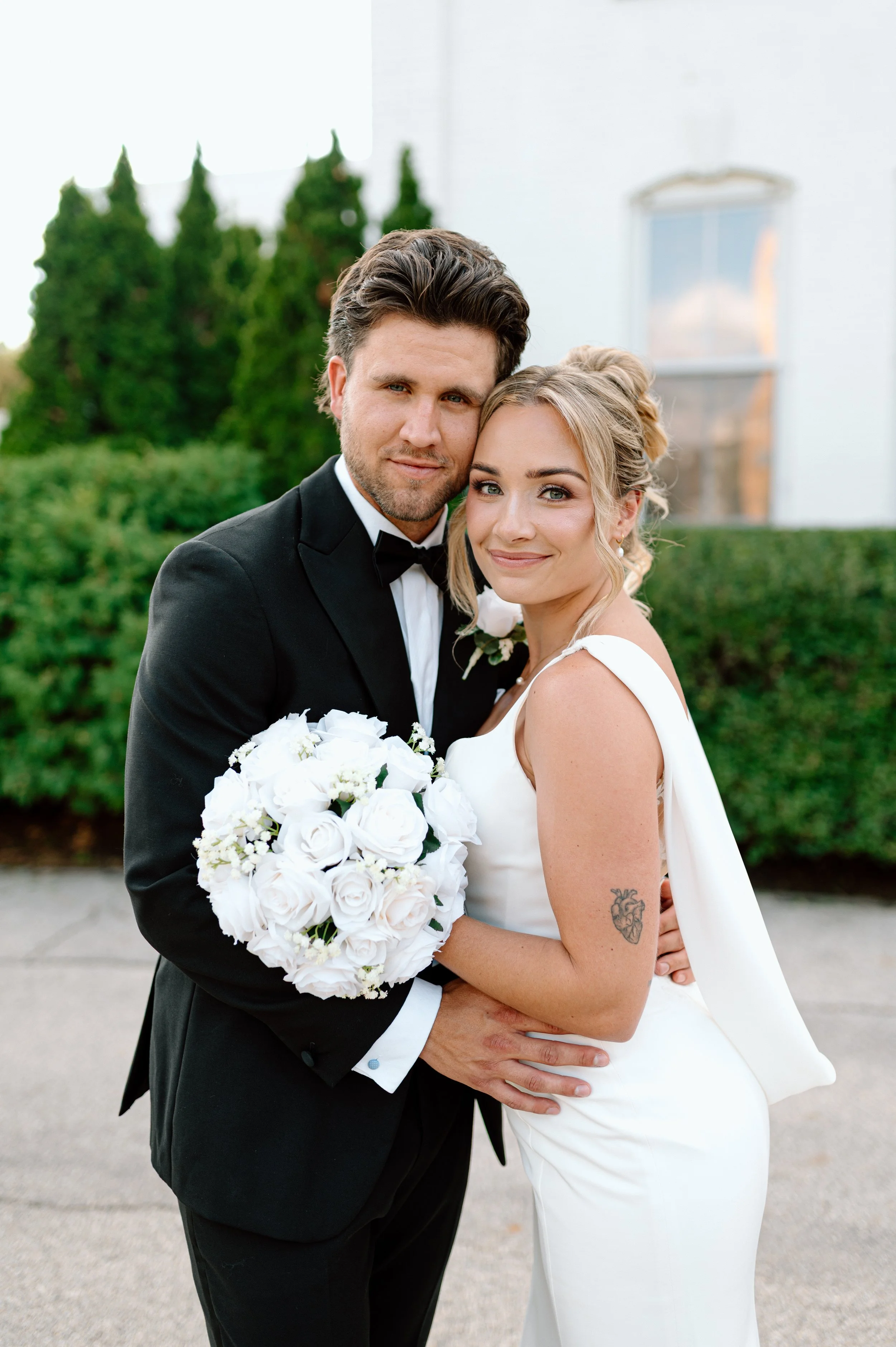 A bride and groom standing close together outside, smiling at the camera. The groom is wearing a black tuxedo with a bow tie, and the bride is in a white wedding dress with her hair styled in an updo. She's holding a bouquet of white roses, and has a tattoo of a heart on her arm.