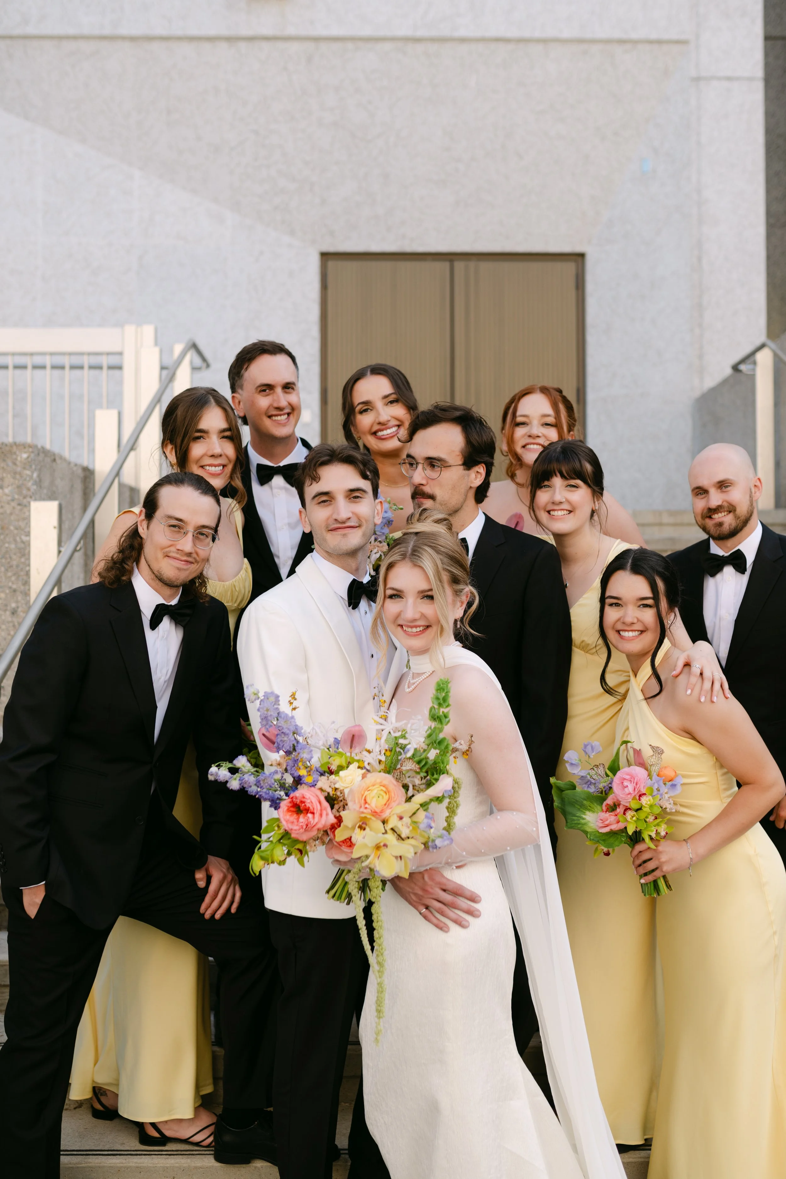 A wedding party with a bride in a white dress holding a bouquet of flowers, surrounded by bridesmaids in yellow dresses and groomsmen in black tuxedos, standing on stairs outside a building.