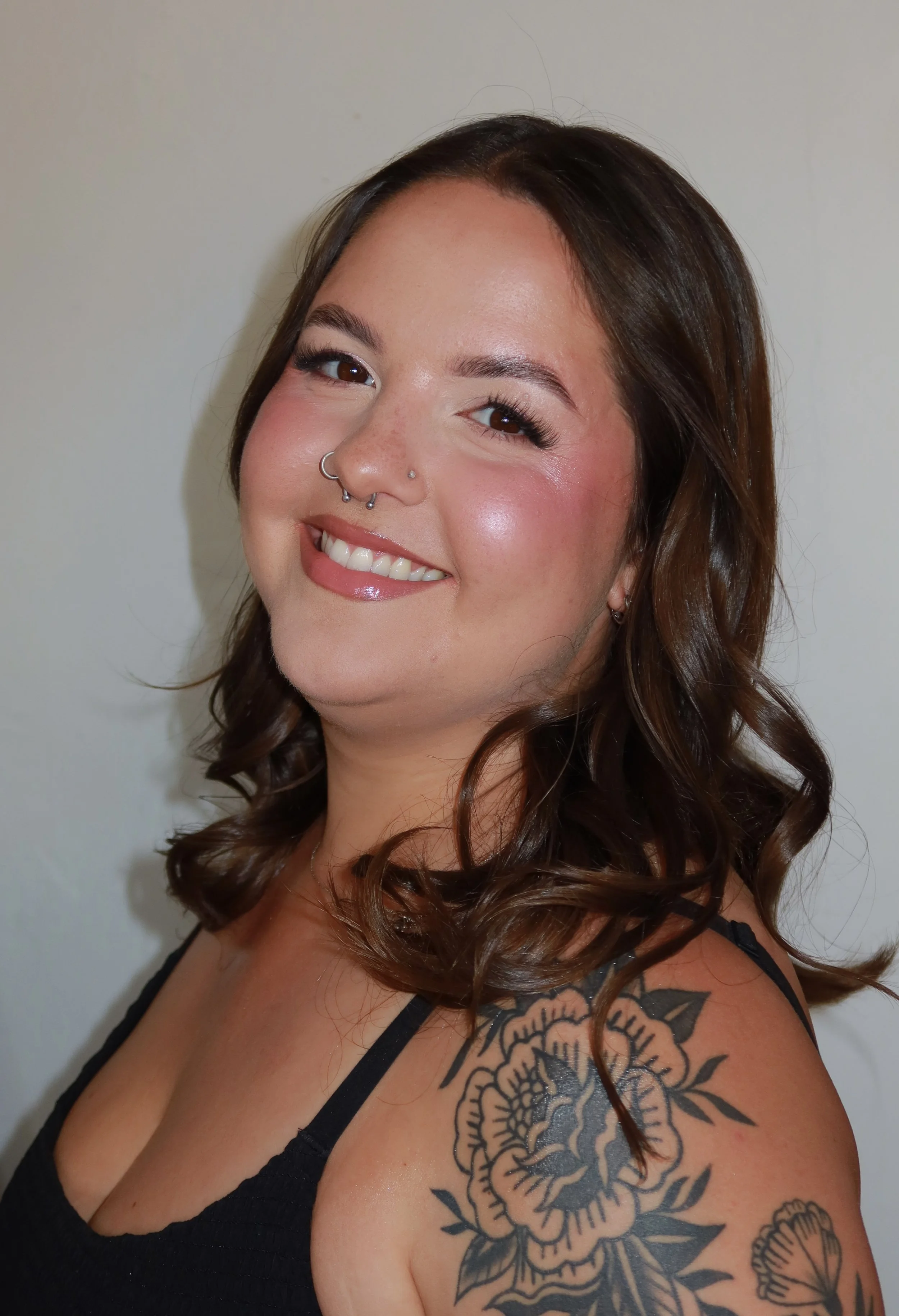 A woman with long brown hair styled in loose curls, smiling, with multiple facial piercings, wearing a black spaghetti strap top, showing a large floral tattoo on her shoulder with roses and a butterfly.