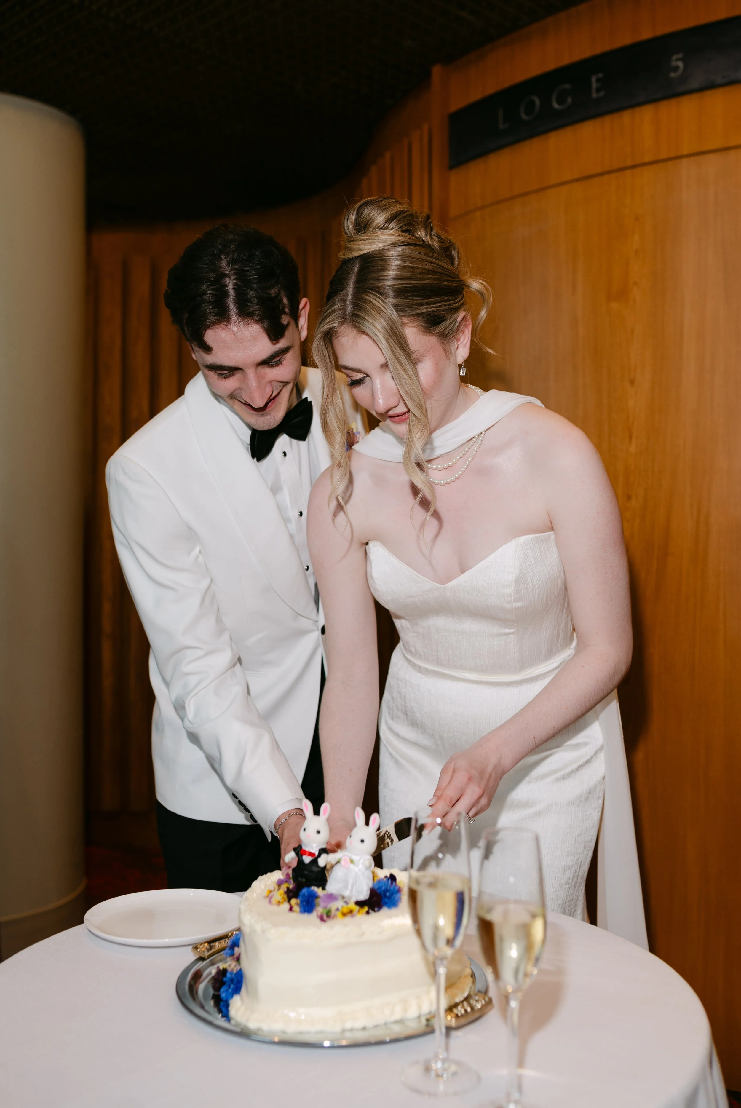 A bride and groom in wedding attire cutting a wedding cake decorated with bunny toppers and colorful flowers at a reception table.
