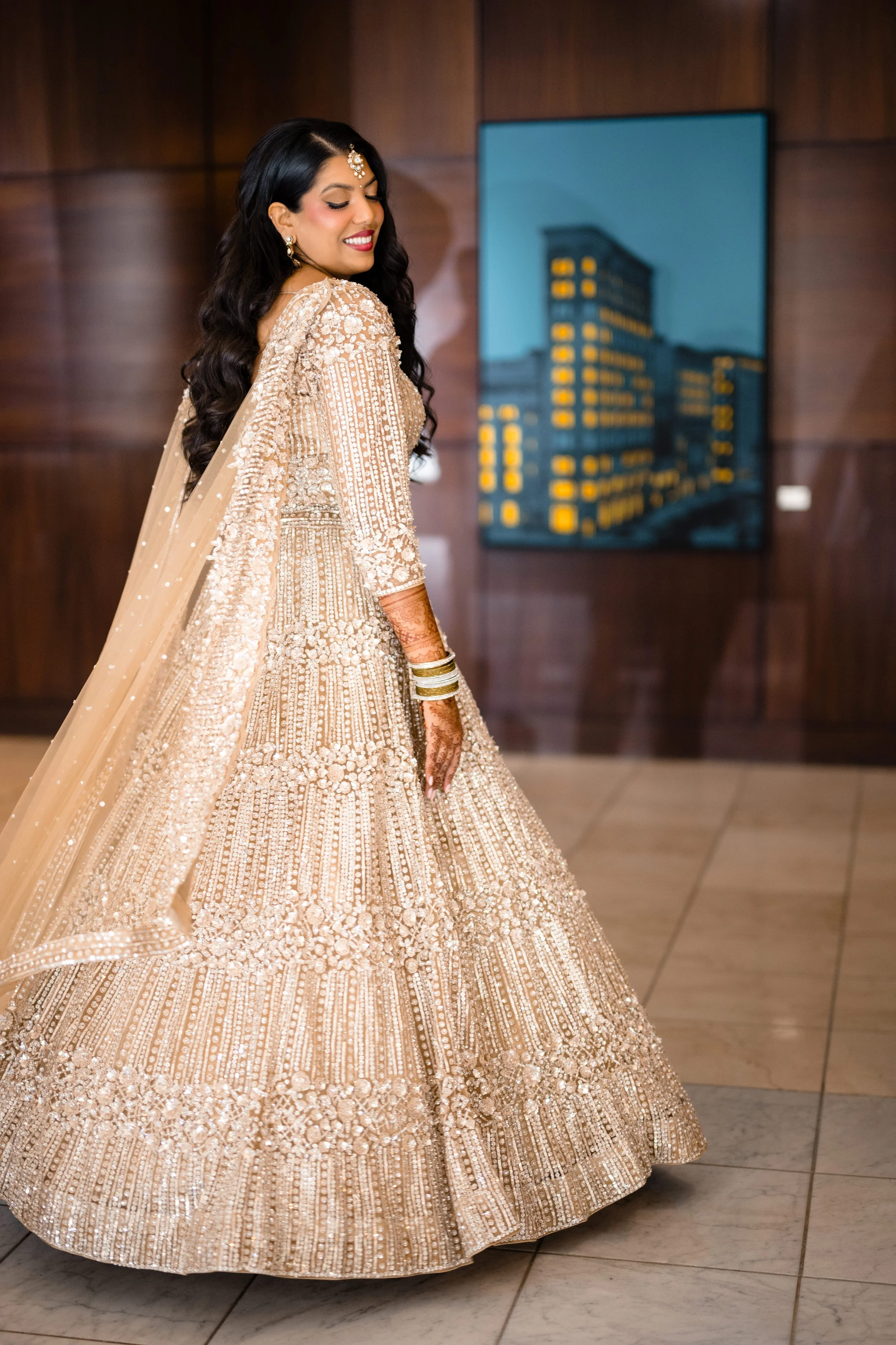 A woman dressed in an ornate, traditional South Asian bridal outfit with intricate embroidery, jewelry, and henna on her hand. She is smiling and turning to the side, standing in an indoor setting with wooden paneling and a cityscape painting in the 