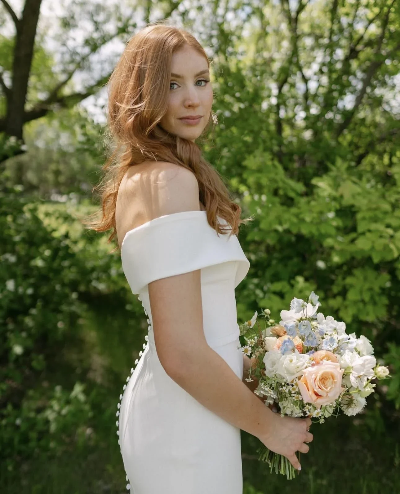 A woman in a white dress holding a bouquet of flowers standing outdoors with green trees in the background.