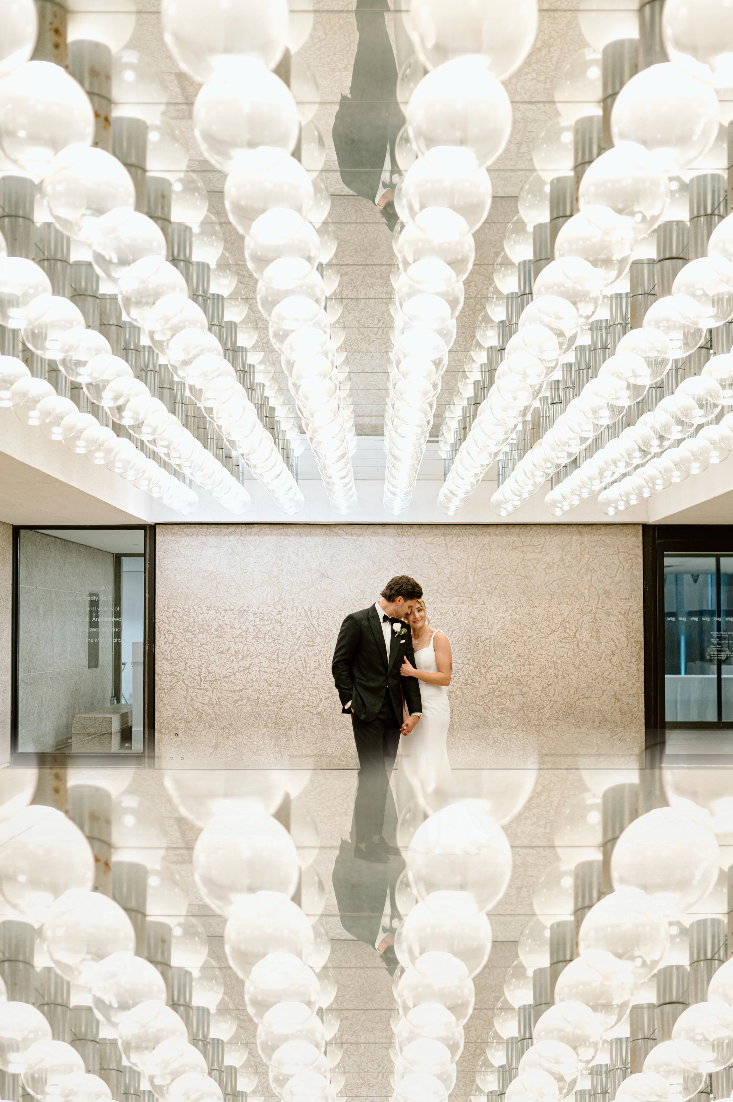 A bride and groom standing closely together inside a modern building, with an elaborate ceiling made of numerous white spherical pendant lights hanging in rows above them.