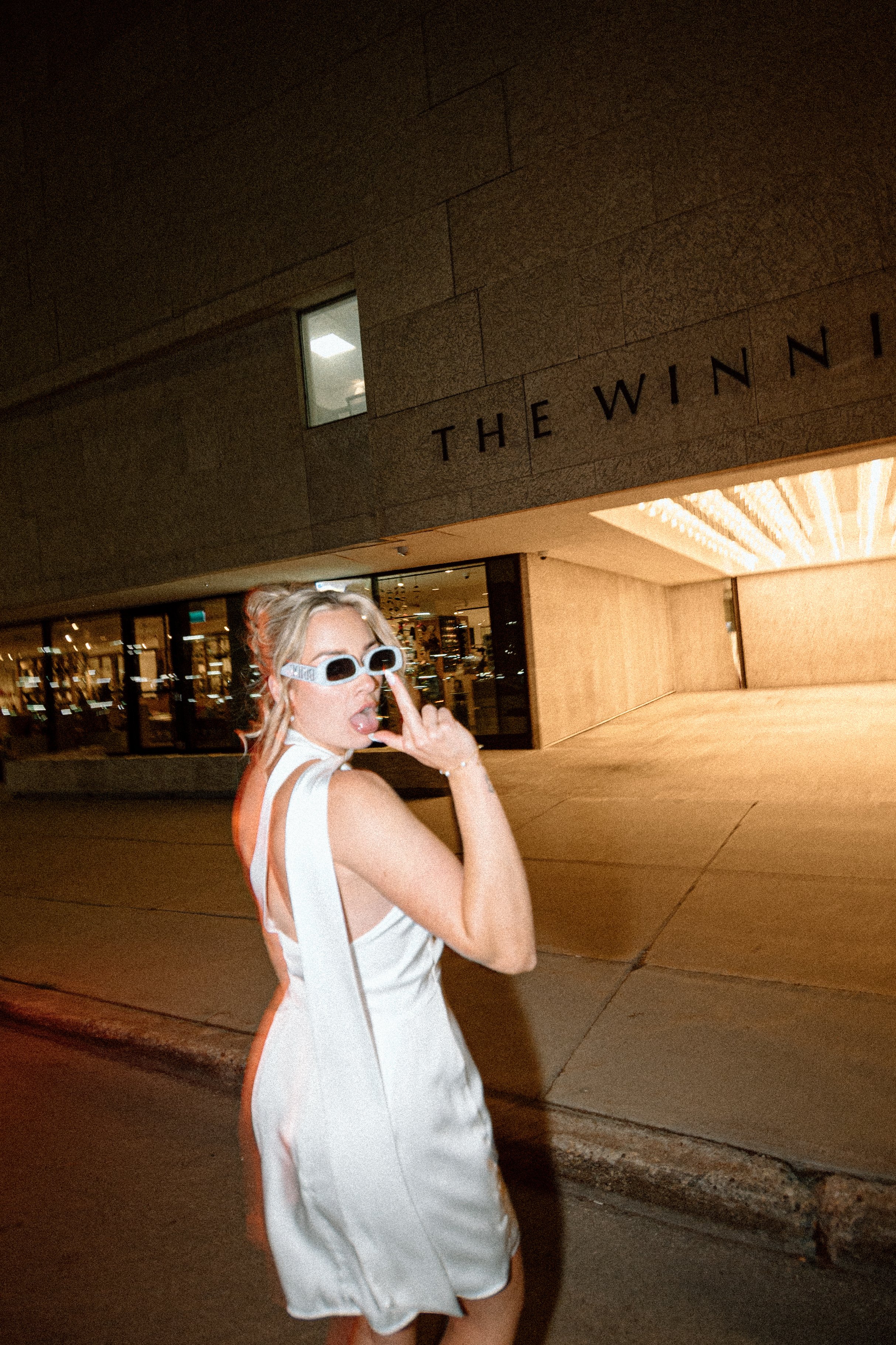 A woman in a white dress and sunglasses is standing outdoors at night in front of a building with the sign 'The Wynn' on it. She is touching her sunglasses with her right hand and sticking out her tongue.