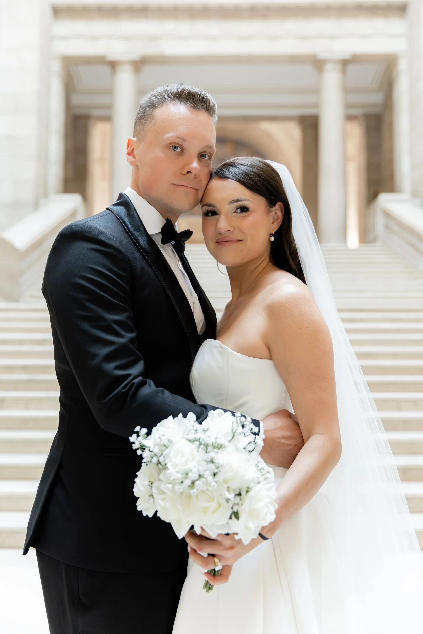 A bride and groom standing close together at a wedding, smiling and looking at the camera inside a grand staircase hall with columns.