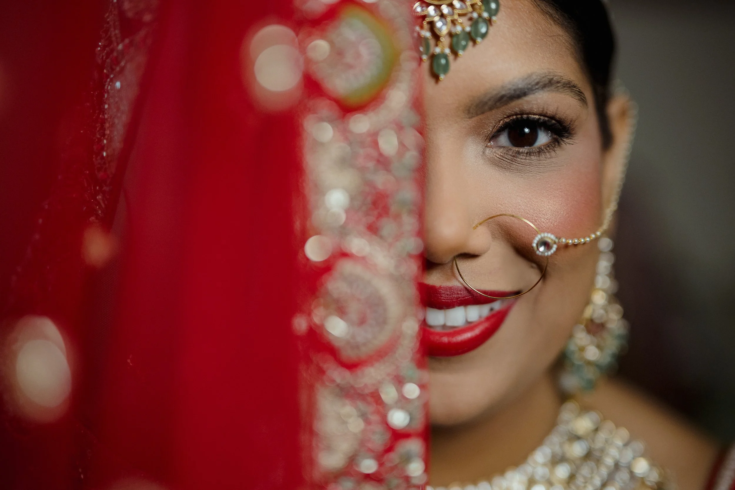 Close-up of a woman wearing traditional jewelry and makeup, partially obscured by a red fabric or veil with sparkling embellishments.