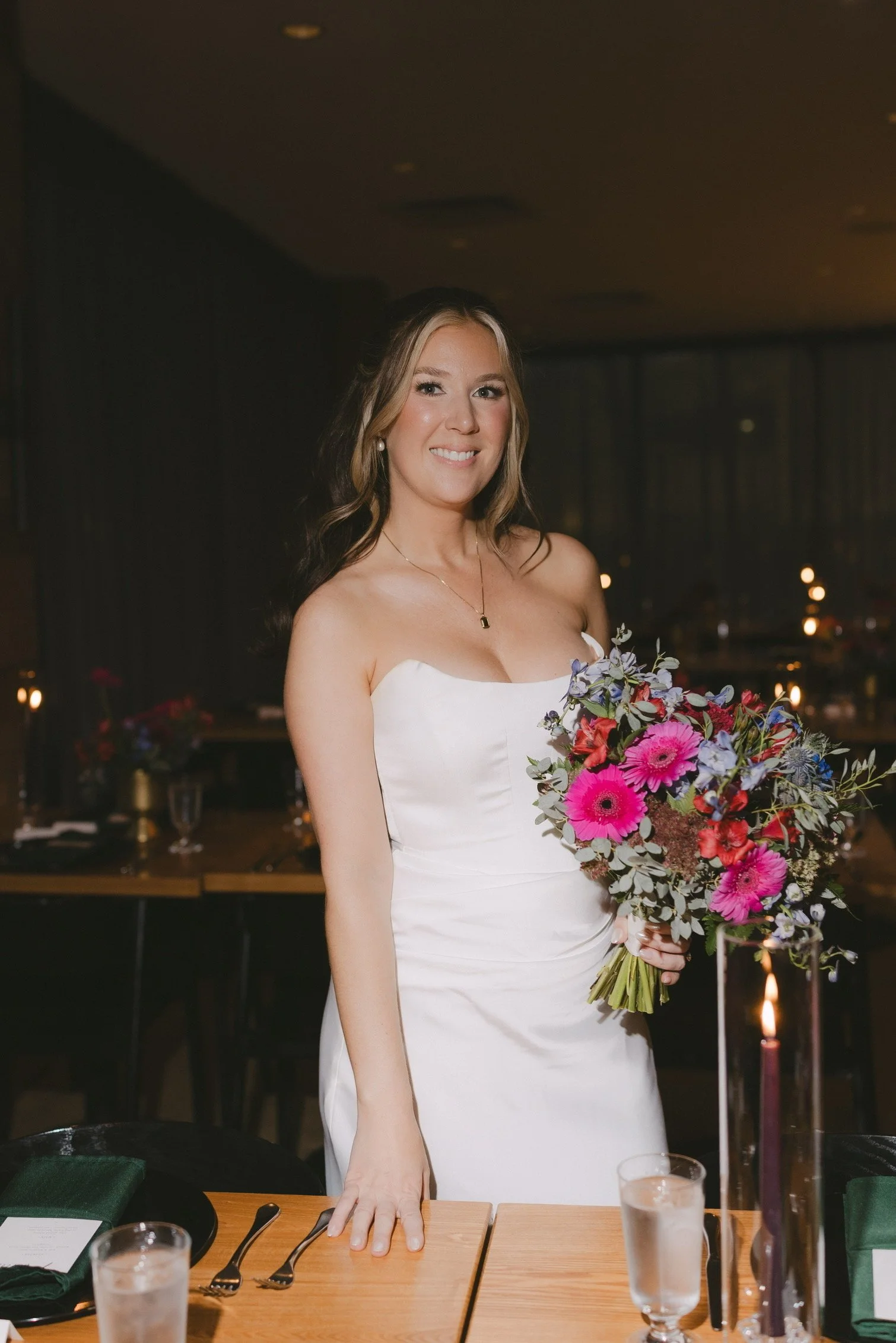 A woman in a white strapless dress holding a bouquet of colorful flowers, smiling at a dinner table in a dimly lit restaurant.