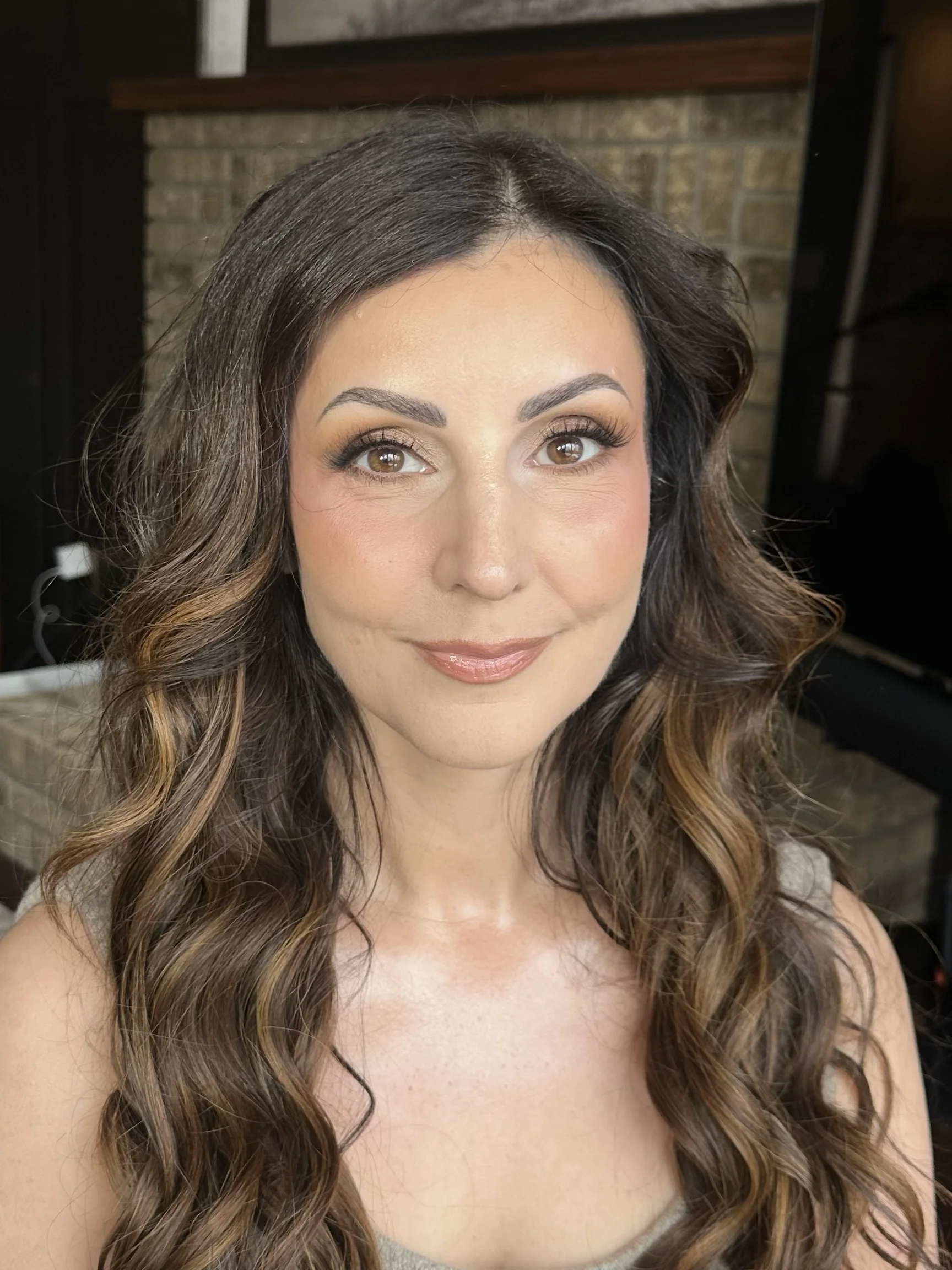 Close-up of a woman with long, wavy brunette hair, light makeup, and a warm smile, sitting indoors with a brick wall and a mirror in the background.
