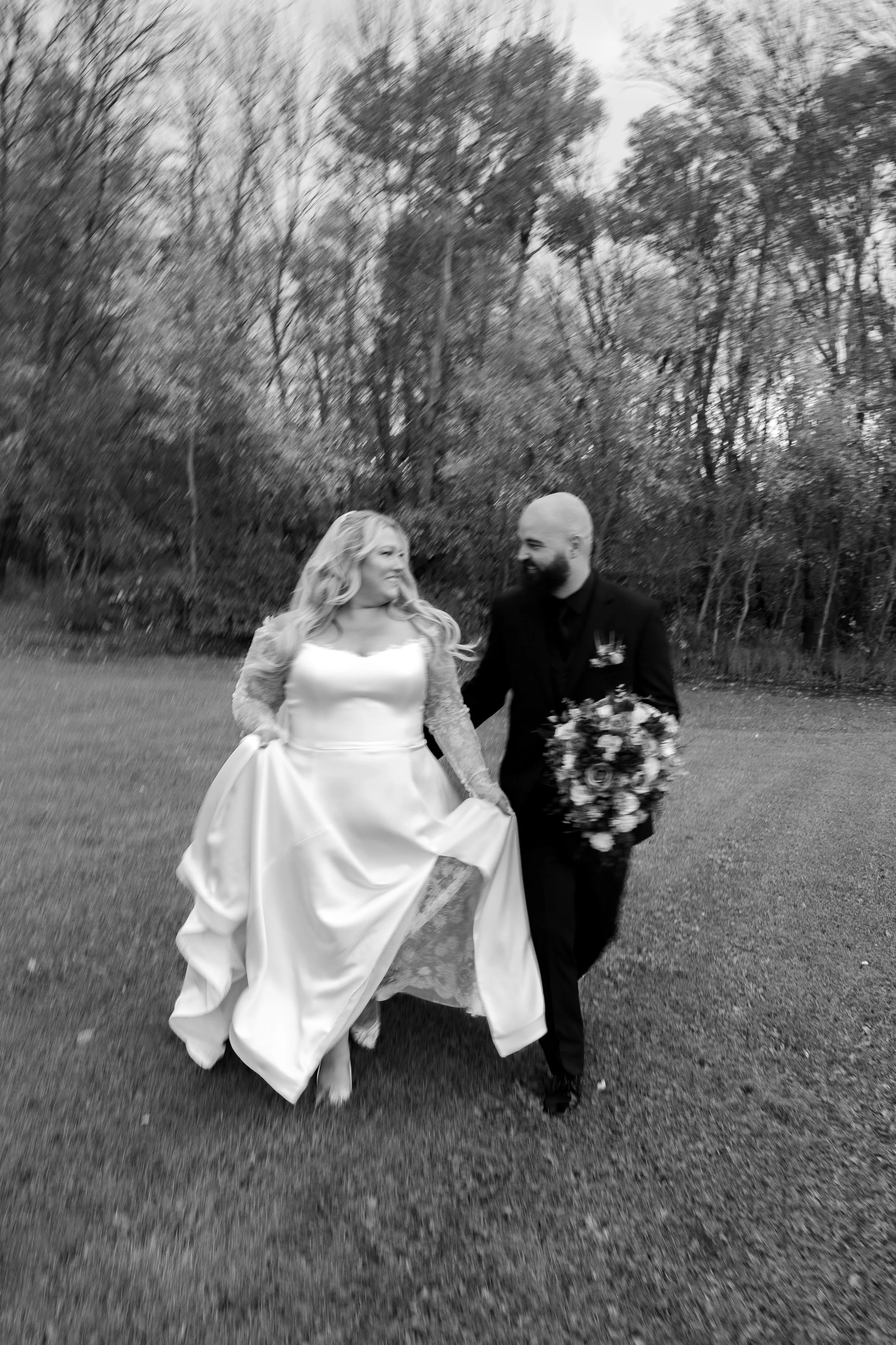 A bride and groom walking outdoors on a grassy field with bare trees in the background, smiling at each other.