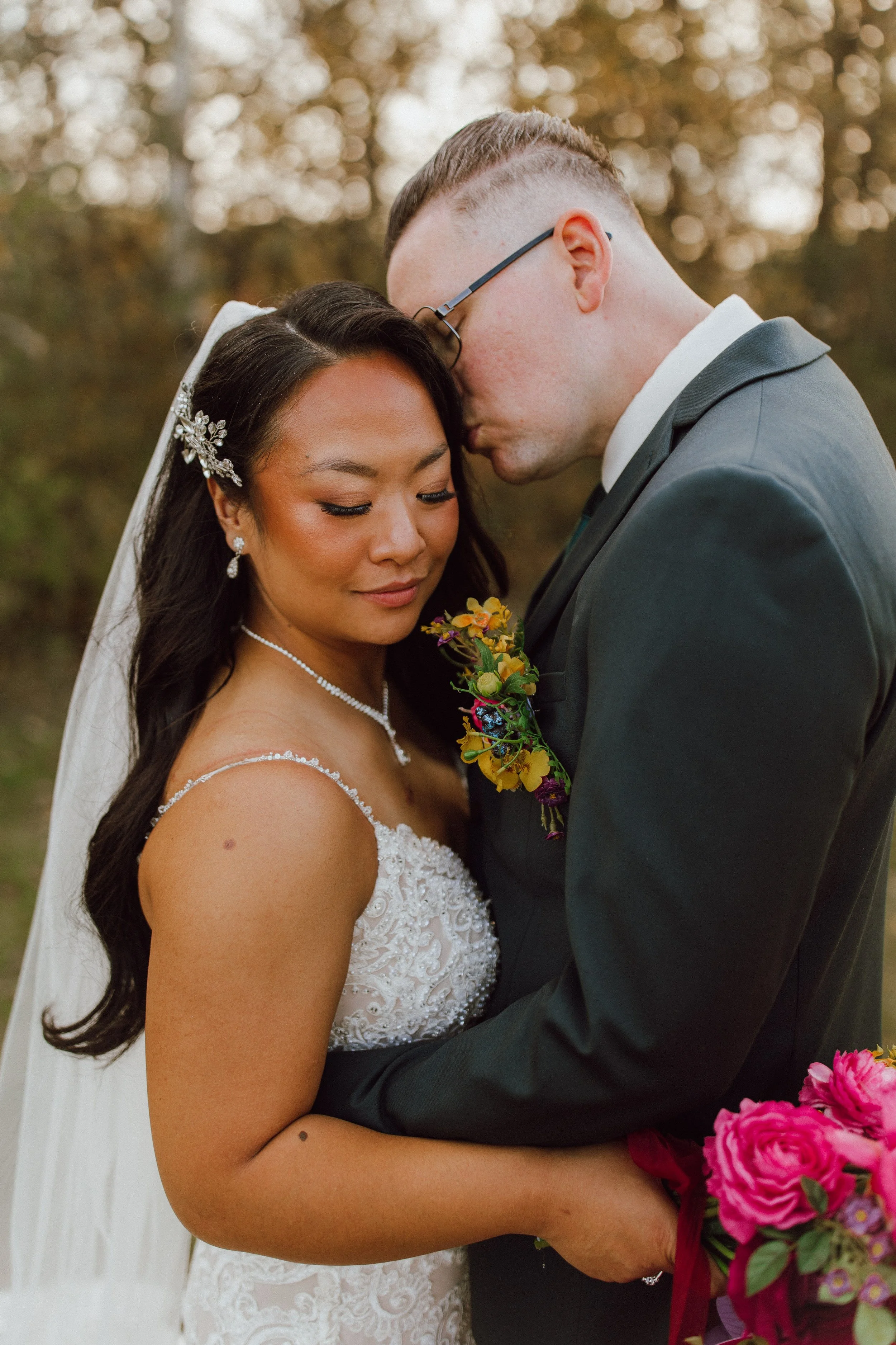 A bride and groom embracing at their wedding, with #bride wearing a white dress with lace details, floral hair accessory, and jewelry, and groom in a gray suit and glasses. The bride holds a colorful bouquet of flowers.