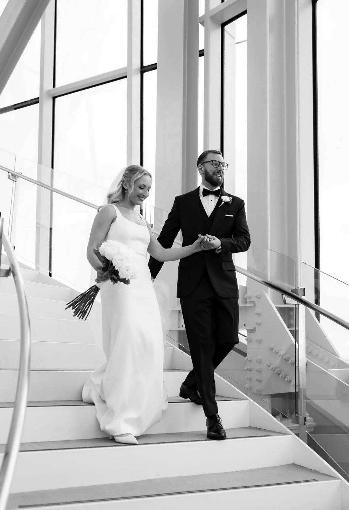 A black and white photo of a bride and groom walking down a staircase in a modern building with large windows. The bride is holding a bouquet of flowers and is wearing a white wedding dress, while the groom is dressed in a tuxedo with a bow tie and glasses.