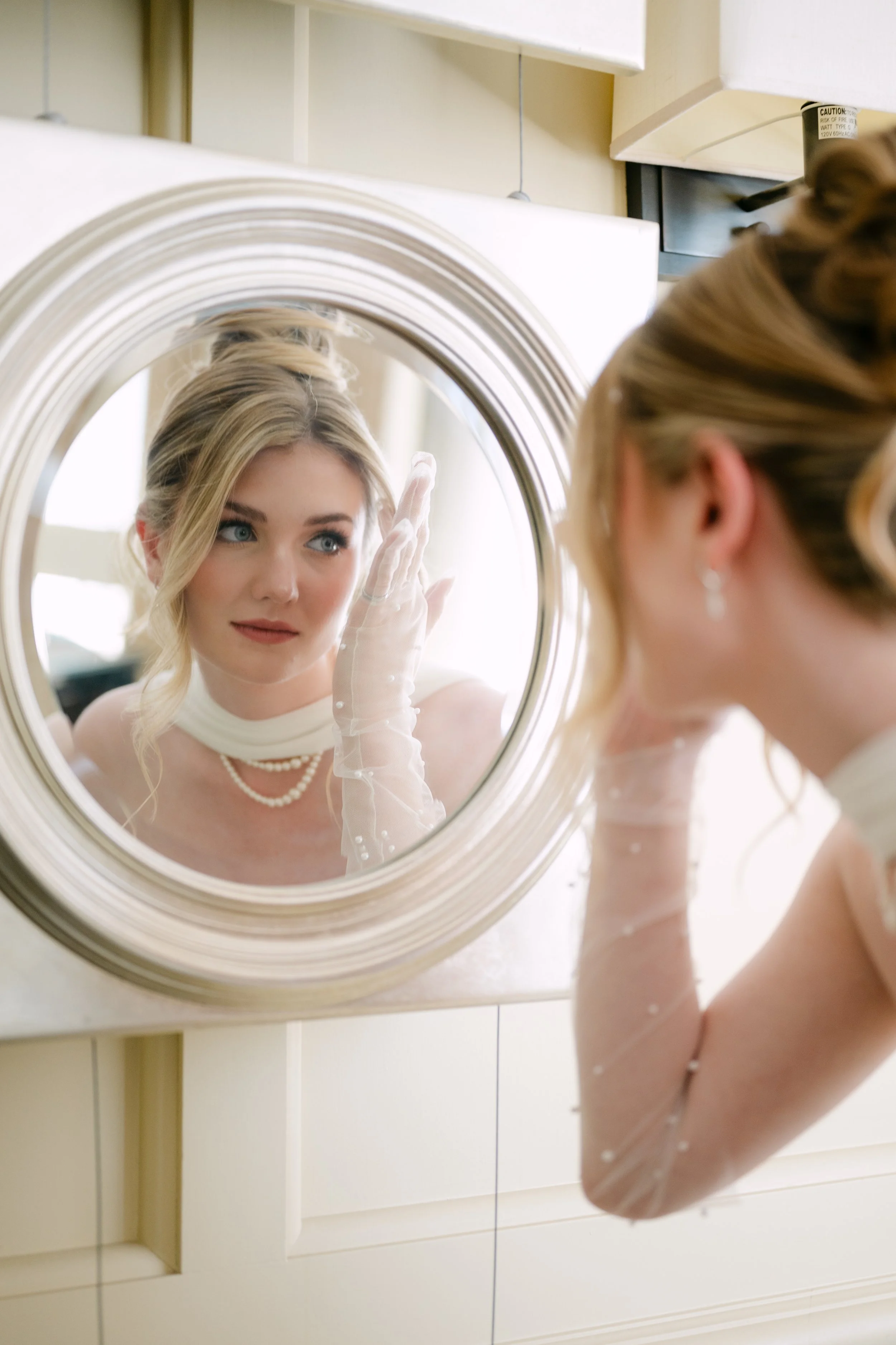 A woman in an elegant dress and pearl necklace looks into a mirror, adjusting her hair or makeup in a bathroom.