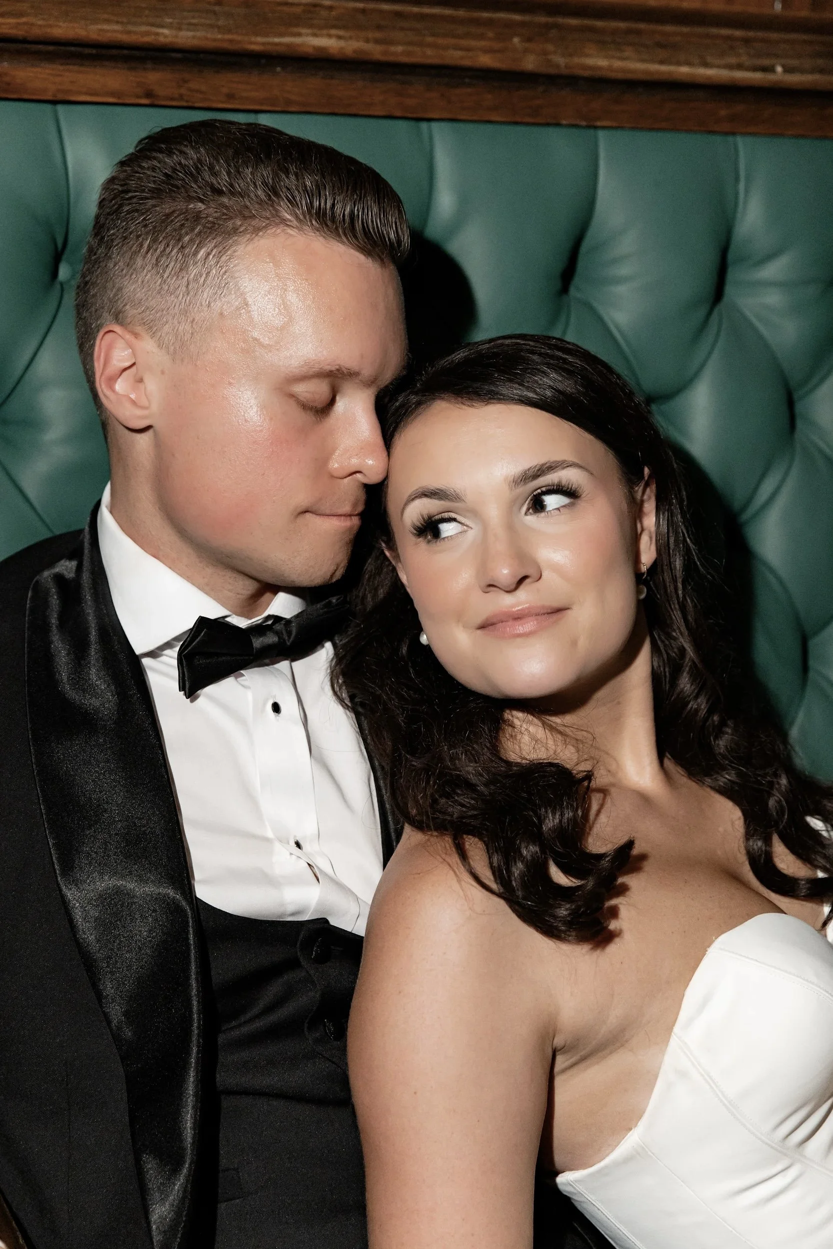 A close-up of a couple in formal attire, with the man kissing the woman's forehead and the woman smiling. The man is wearing a black tuxedo and the woman is wearing a strapless white dress.