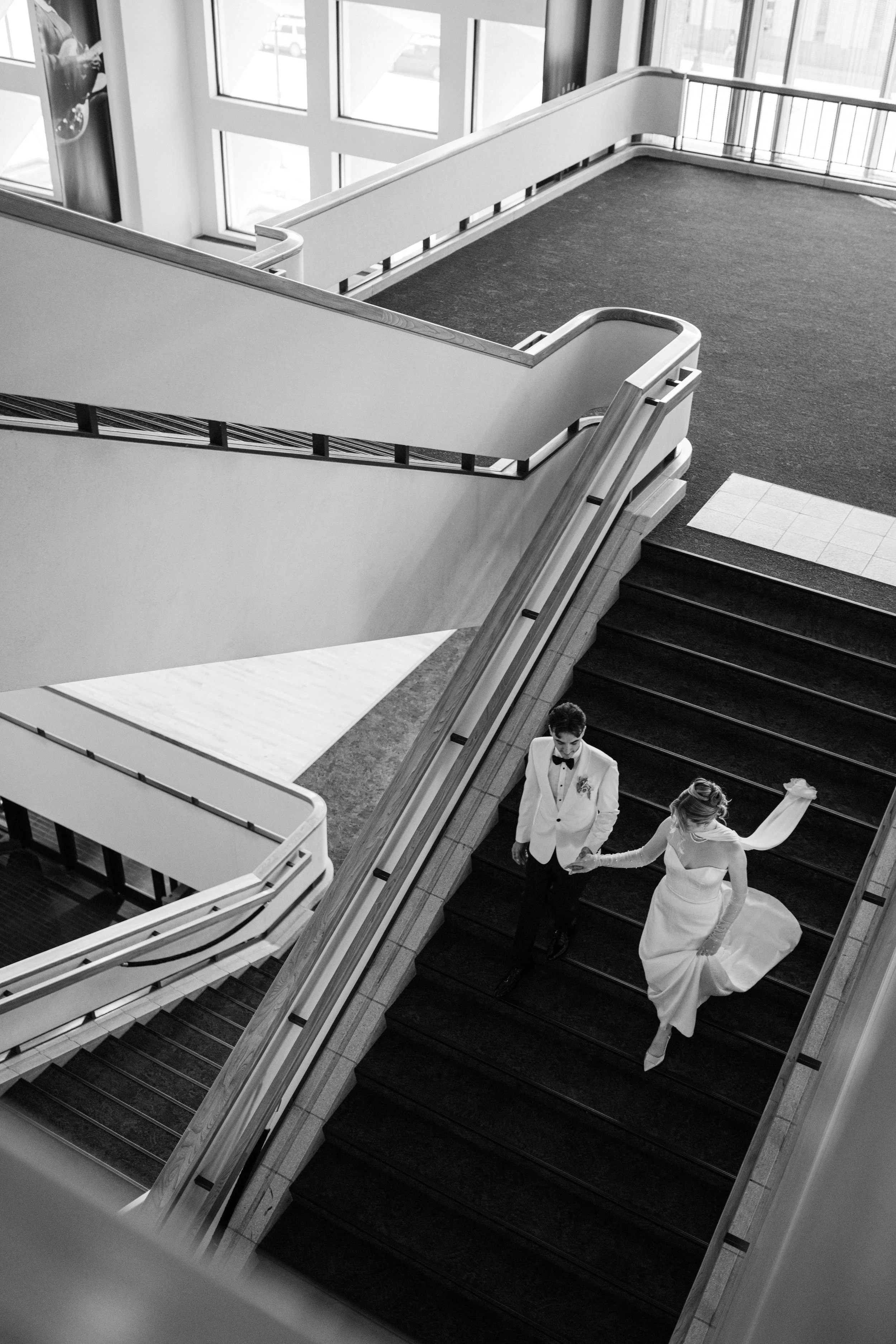 A bride and groom walking down a staircase during their wedding, holding hands, with the bride wearing a white dress and veil, and the groom in a tuxedo. The staircase is indoors with large windows and railing.