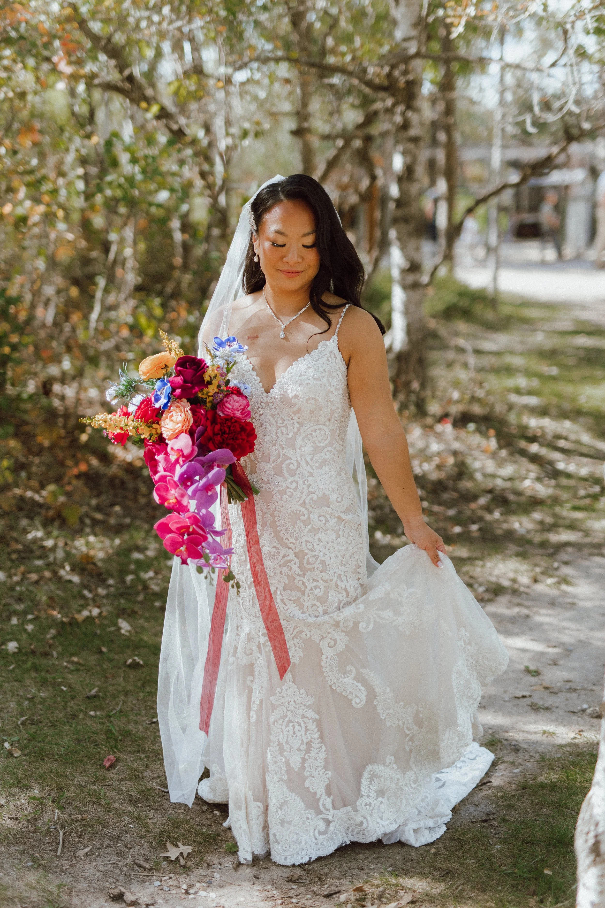 A bride in a white lace wedding dress holding a colorful bouquet of flowers, standing outdoors on a path covered with fallen leaves, surrounded by trees with autumn foliage.
