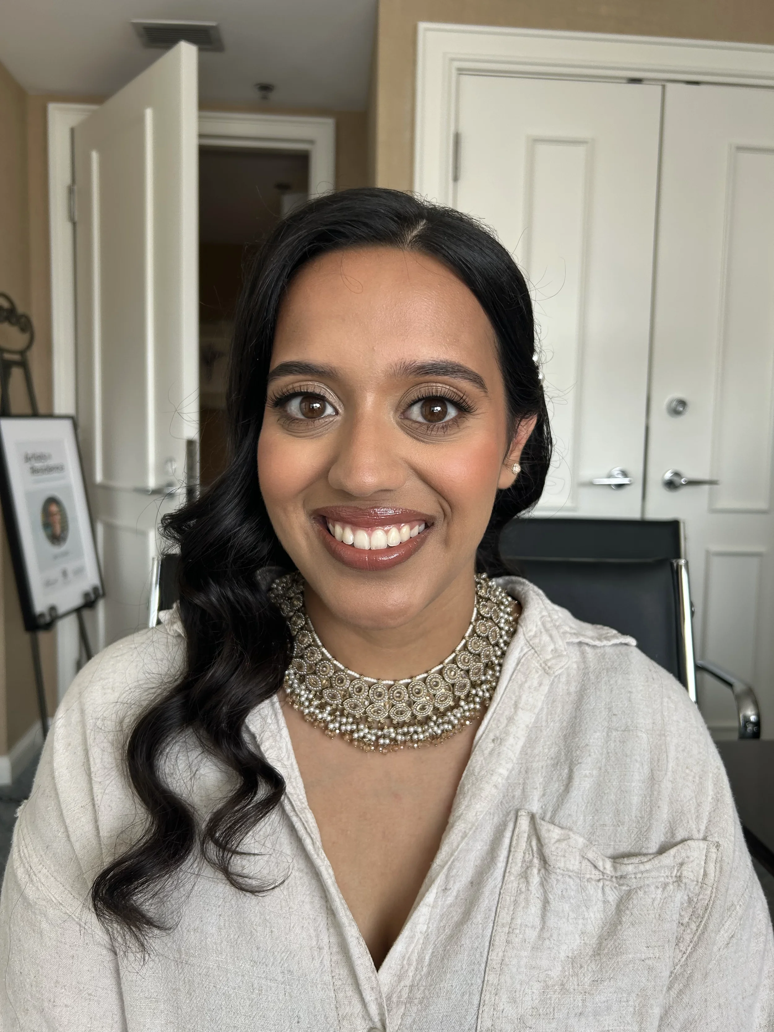 A woman smiling in a hair salon, wearing a white sweater and a statement pearl necklace, with dark wavy hair styled to one side, seated in front of a mirror and salon tools.