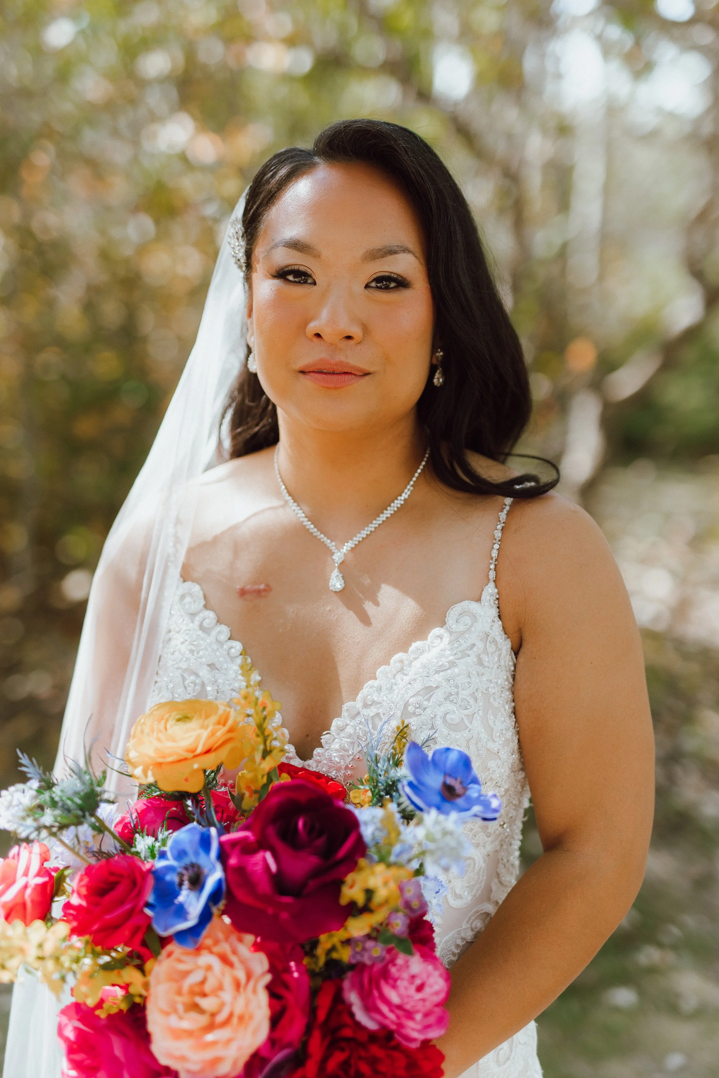 A bride in a white wedding gown holding a colorful bouquet of flowers outdoors with blurred green and brown foliage in the background.