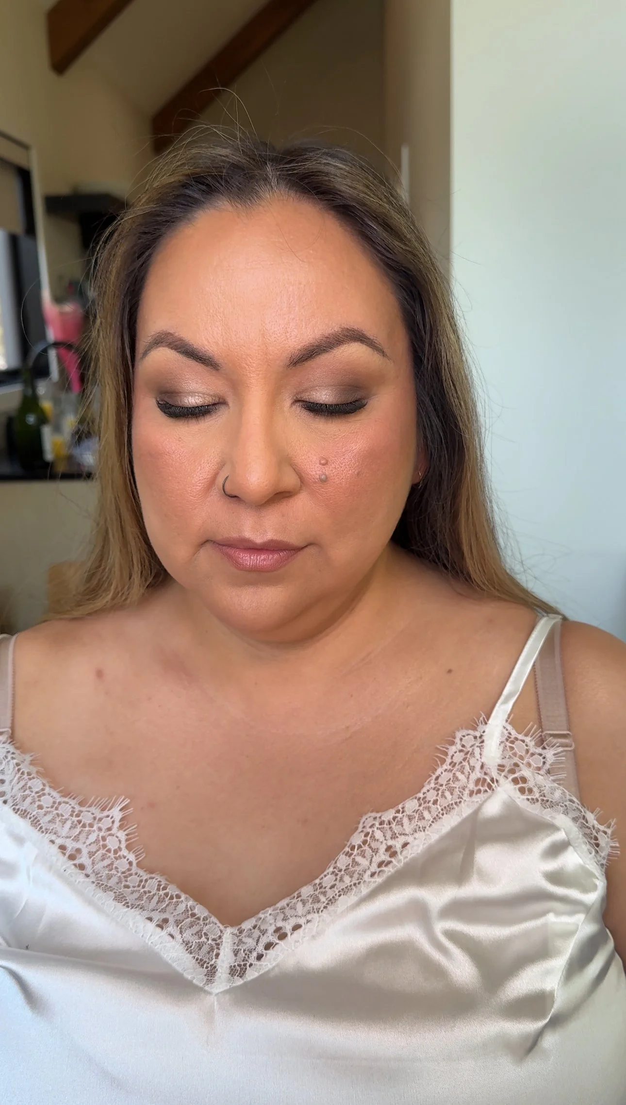 A woman with light skin tone, closed eyes, wearing makeup including eyeshadow and lipstick, a nose ring, and a lace-trimmed satin white top. She is indoors with a blurred background, and her hair is styled loosely.