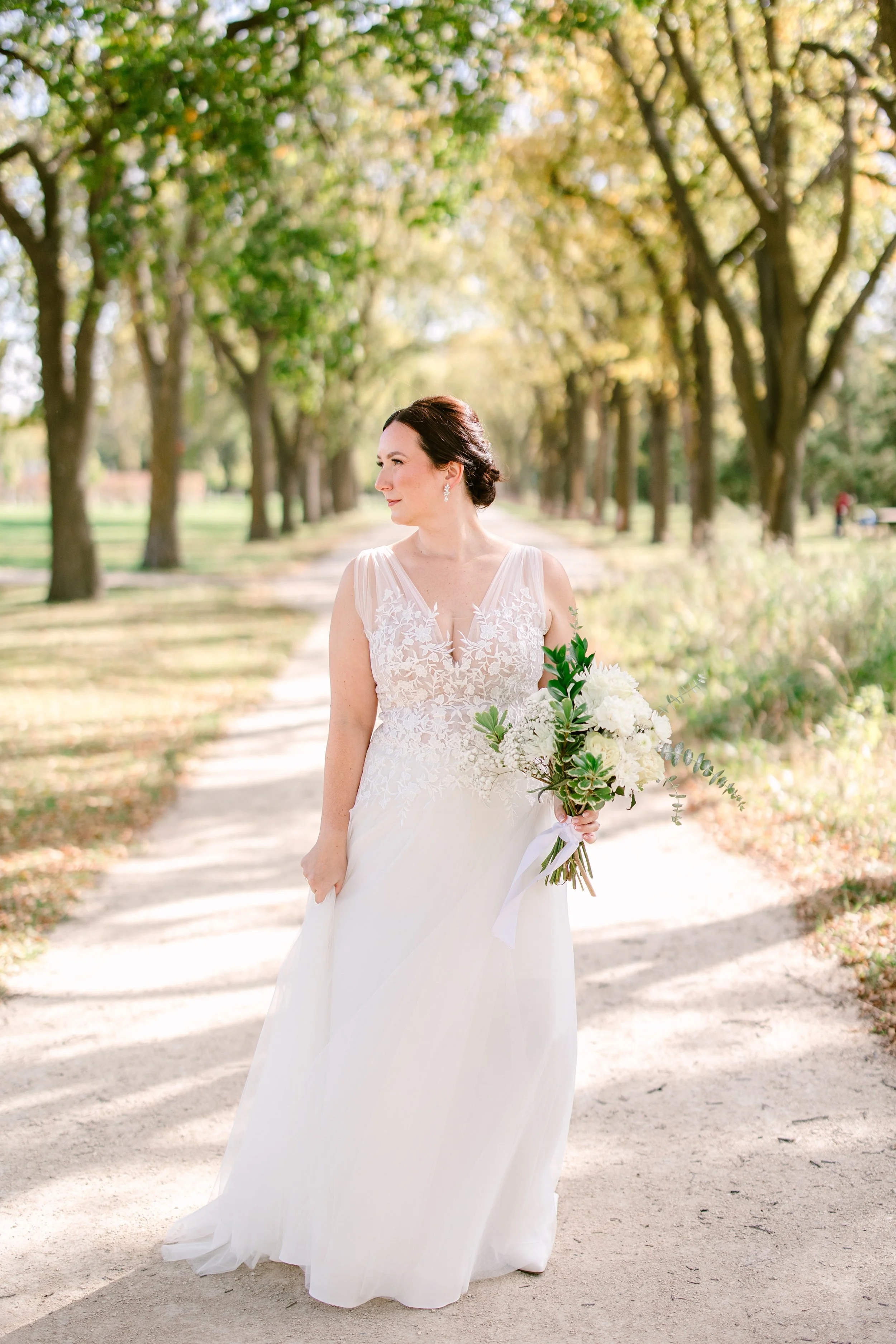 A woman in a wedding dress holding a bouquet of white flowers and greenery on a tree-lined pathway in a park on a sunny day.