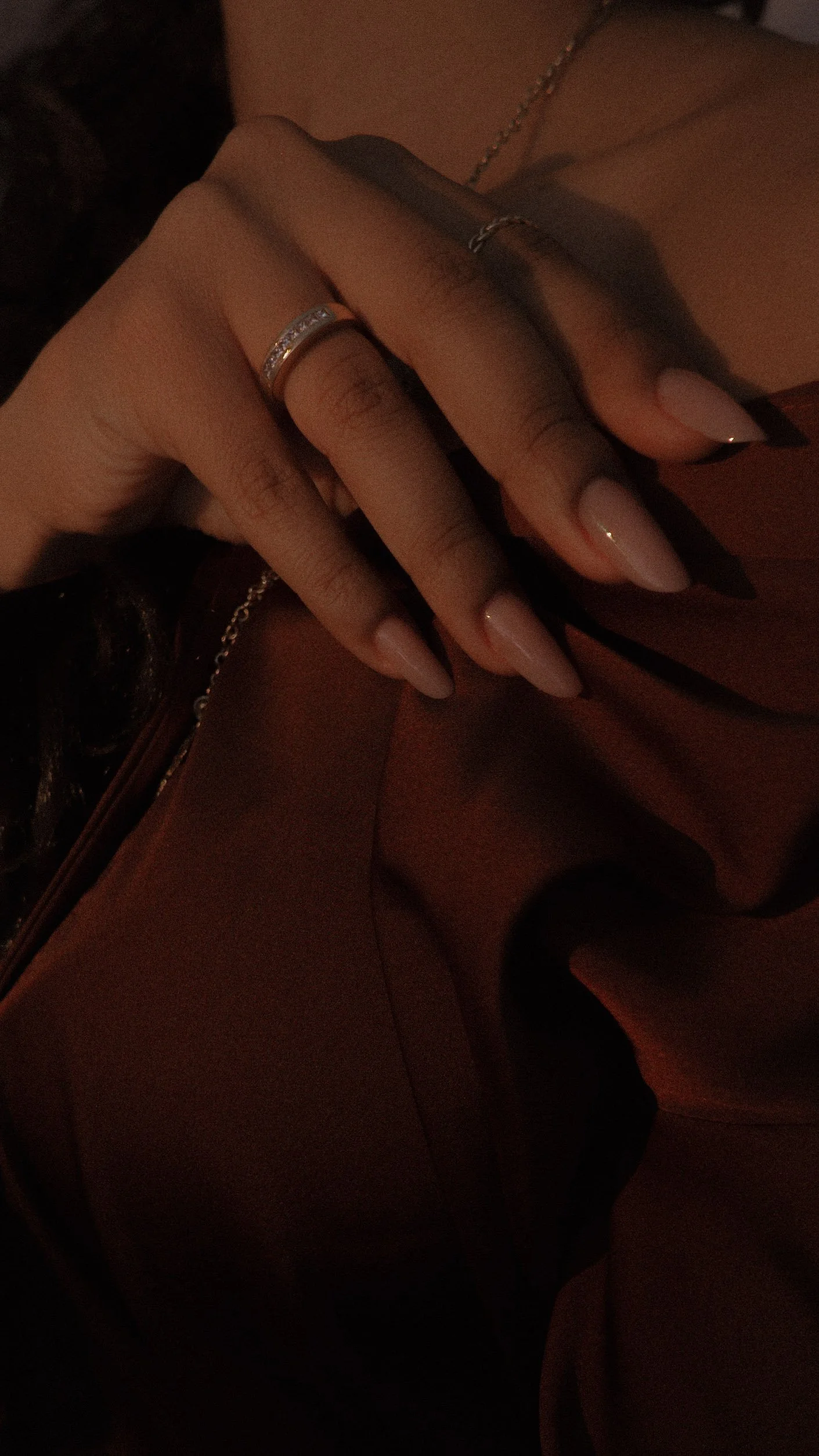 Close-up of a woman's hand resting on her chest, showing manicured nails, a silver ring with stones, and a thin bracelet. She is wearing a chain and a brown garment with a zipper.