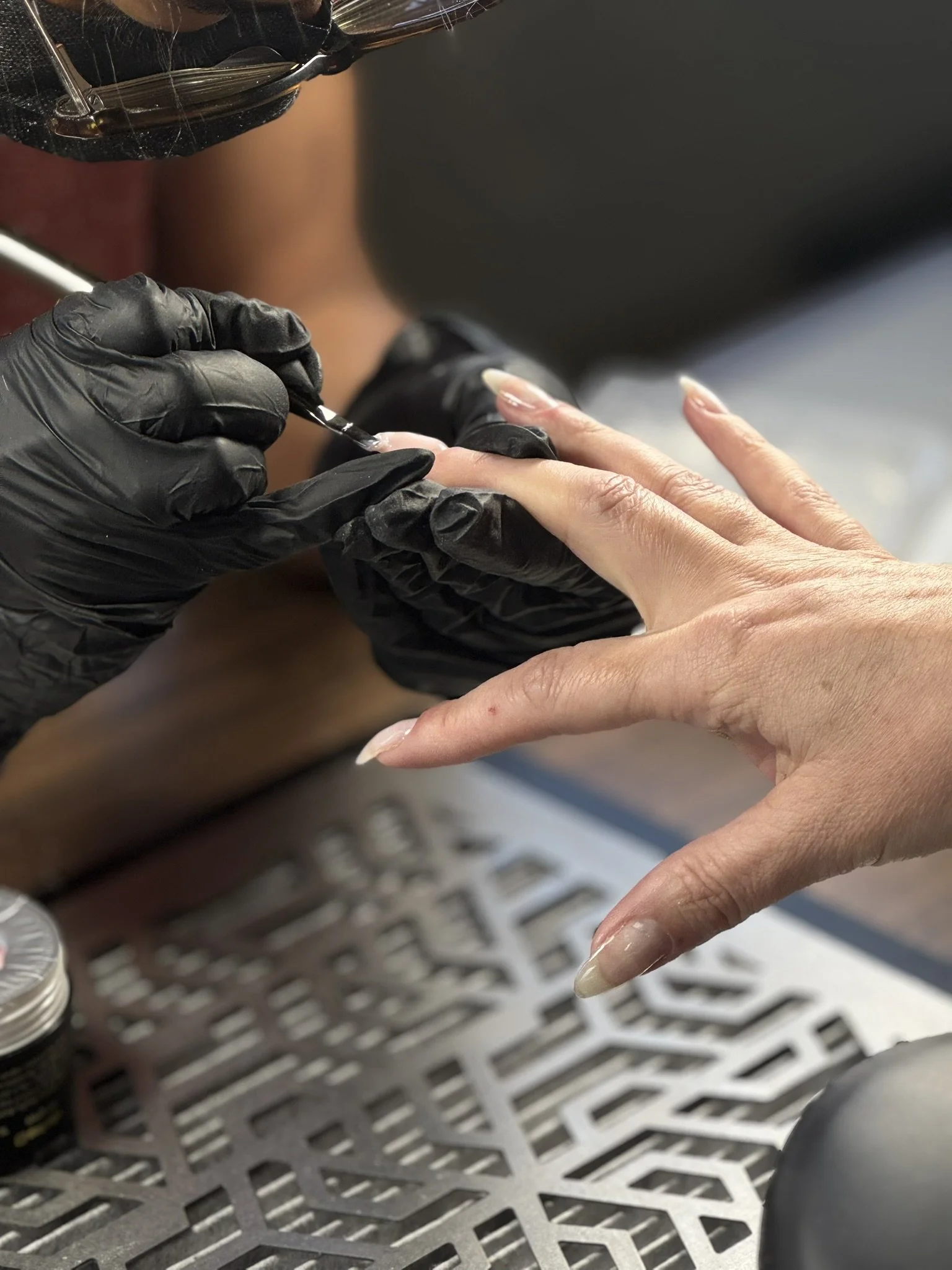 Close-up of a person receiving a manicure with clear nail polish, and the nail technician wearing black gloves applying the polish to the client's fingernails.