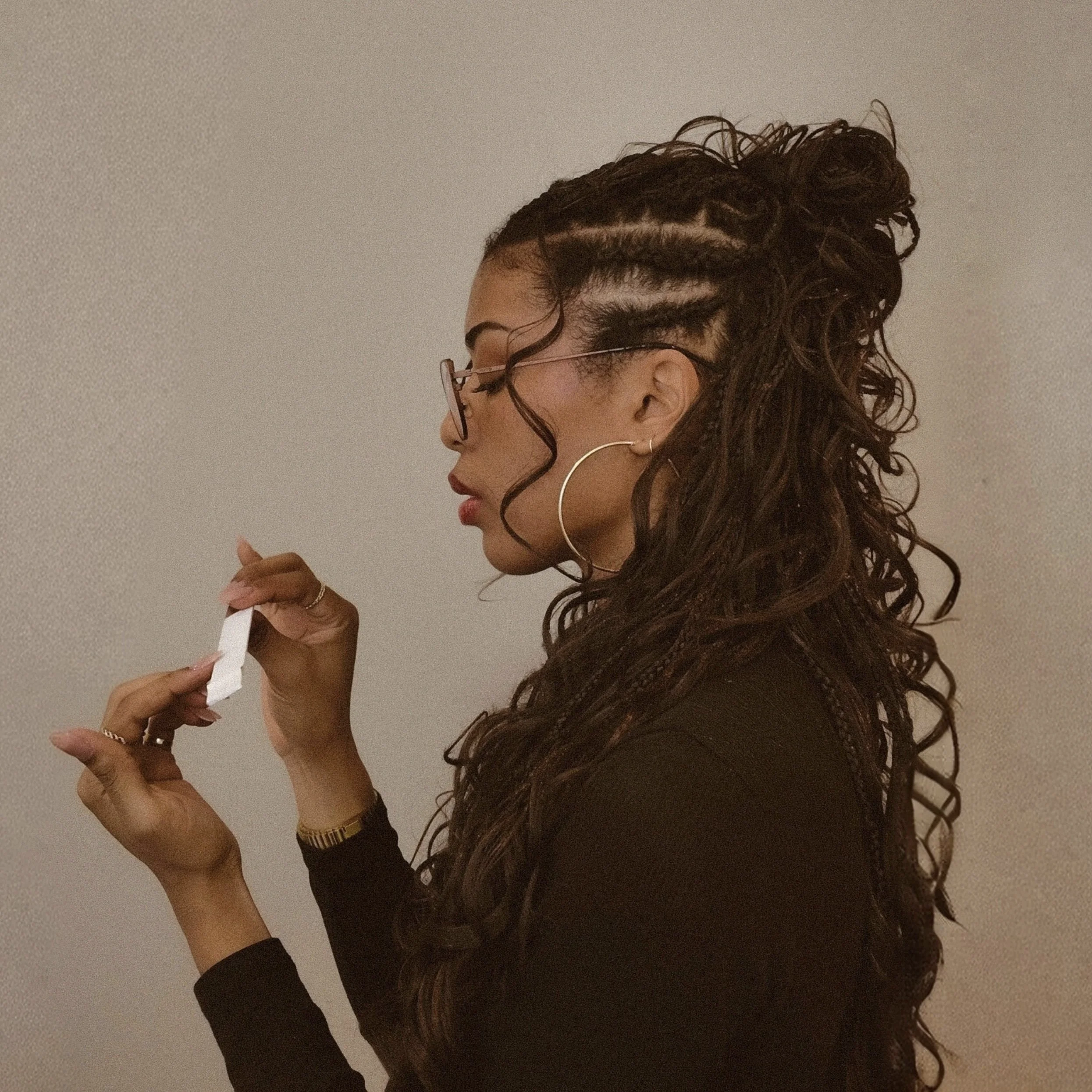 Side view of a woman with long, curly hair in braid style, wearing large hoop earrings, glasses, and a black top, holding and looking at a small object in her hand.