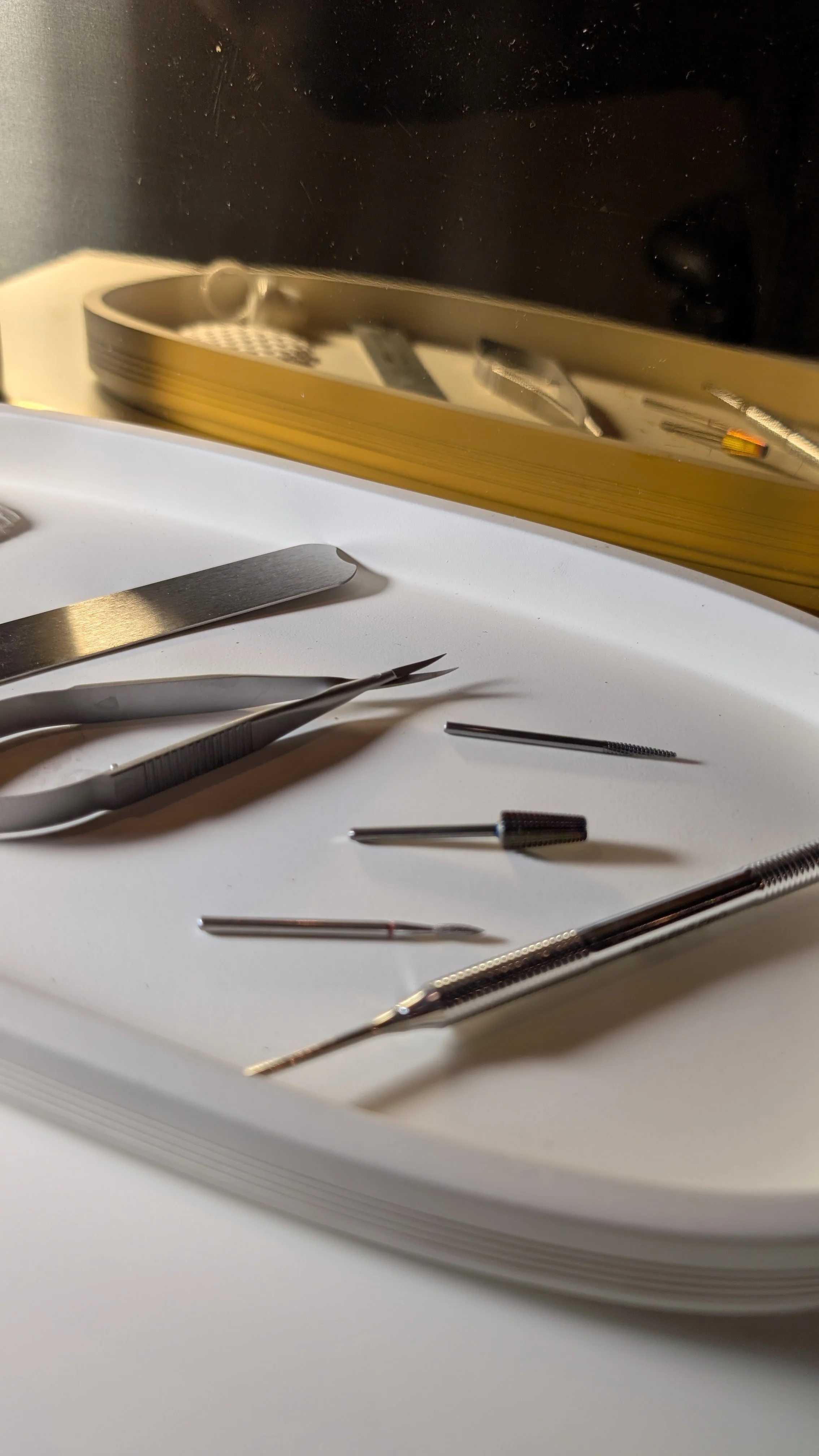 A close-up of various tiny metal tools and tweezers placed on a white surface, with a gold tray and a yellow-edged black object reflected in the background.