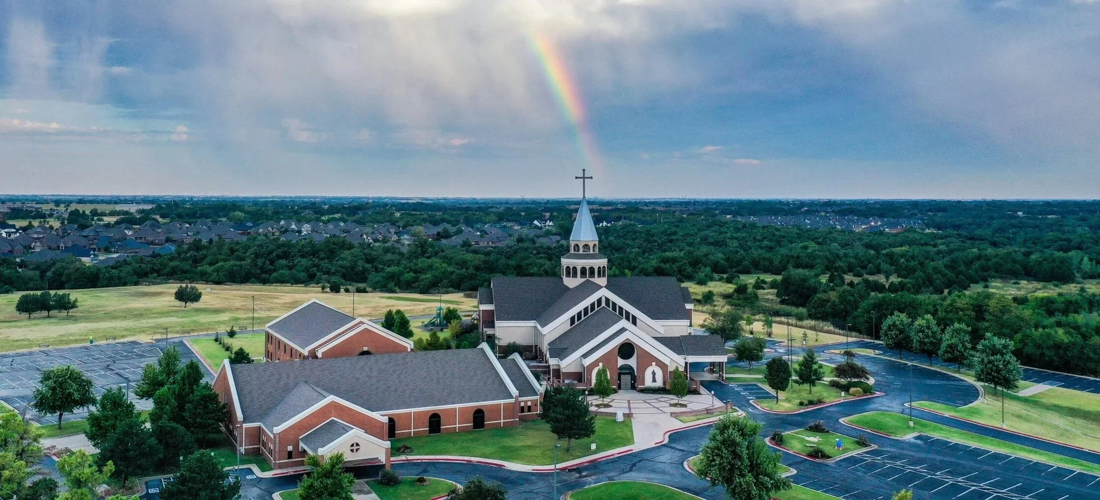 An aerial view of a church with a parking lot, surrounded by green trees and open fields under a cloudy sky with a rainbow.