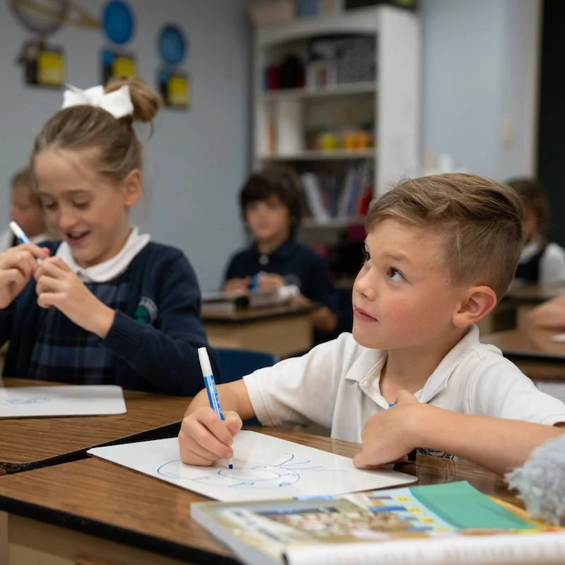 Young boy and girl in classroom, girl laughing as she draws with a marker, boy looking at her, other students in background