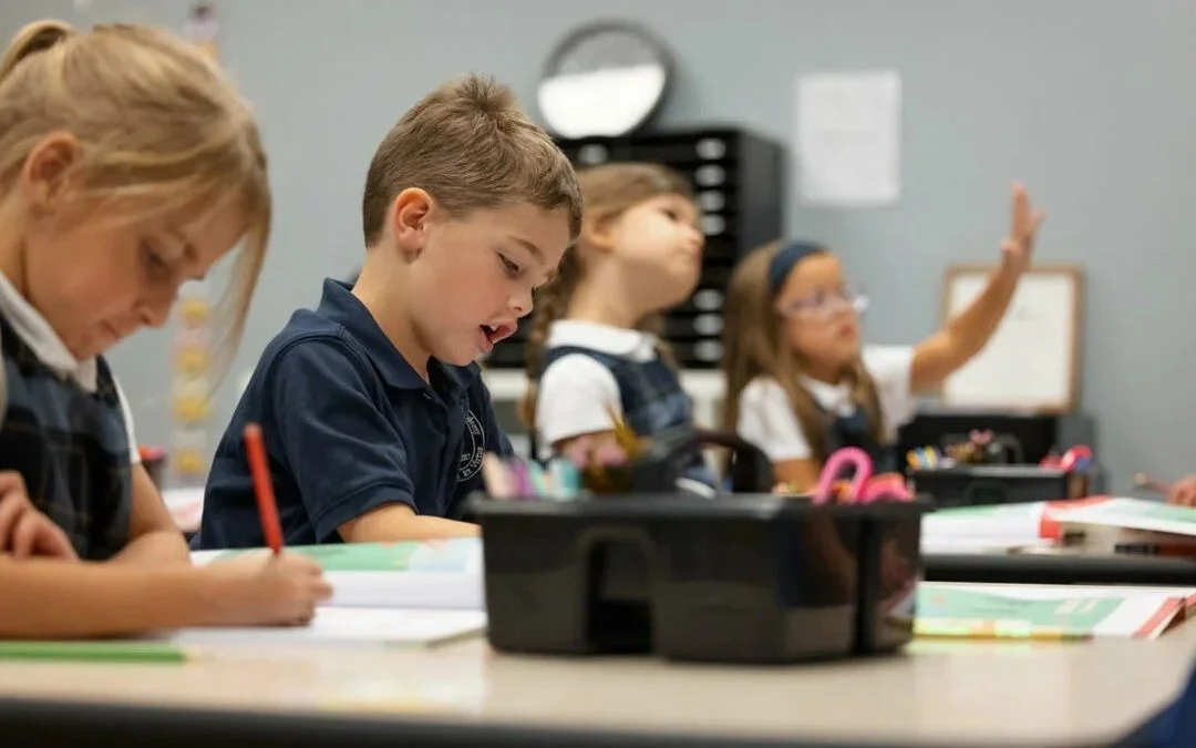 Four elementary school children sitting at desks in a classroom, engaged in learning activities. One girl is raising her hand.
