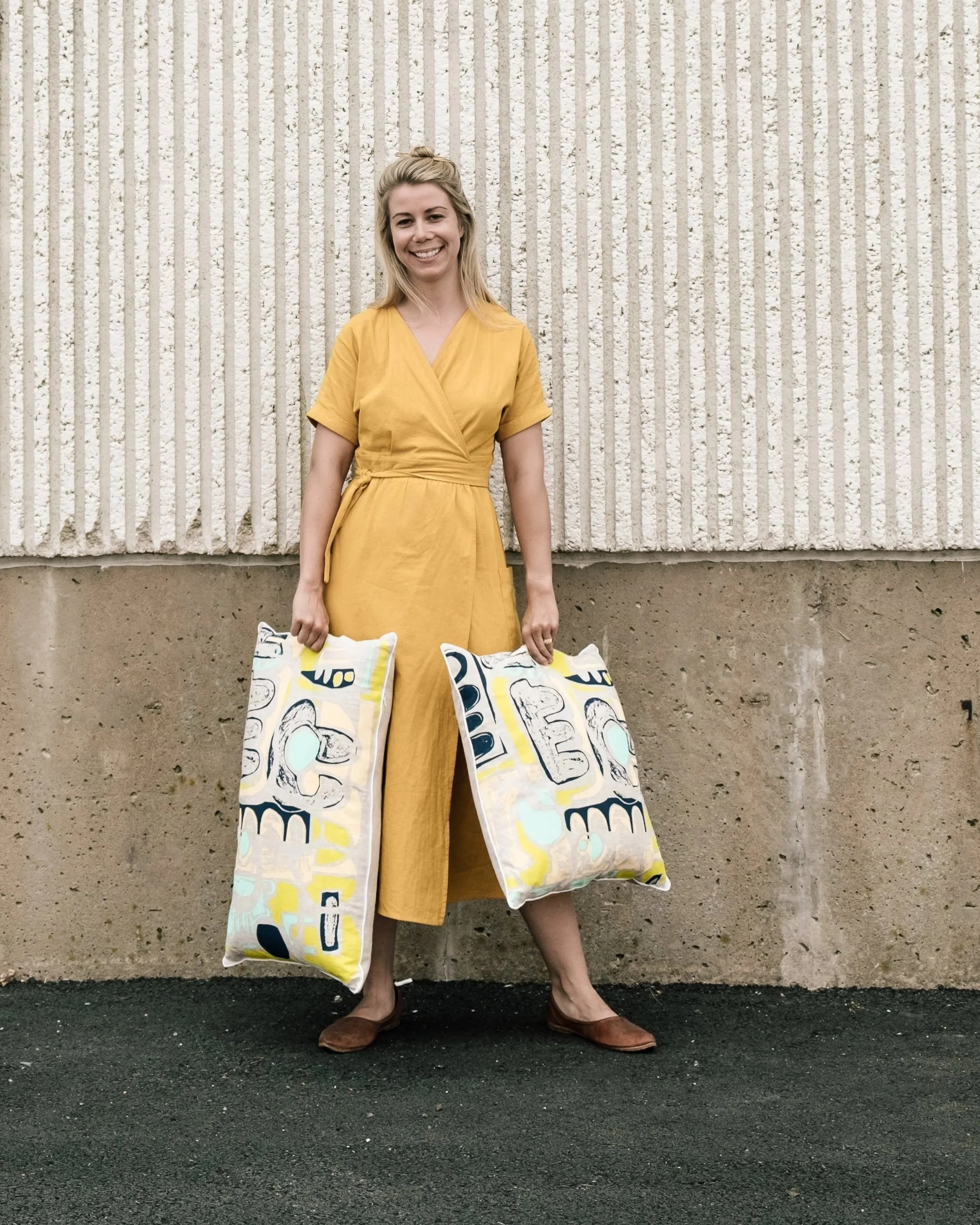 Woman in a yellow wrap dress holding shopping bags with abstract patterns, standing against a textured concrete wall.