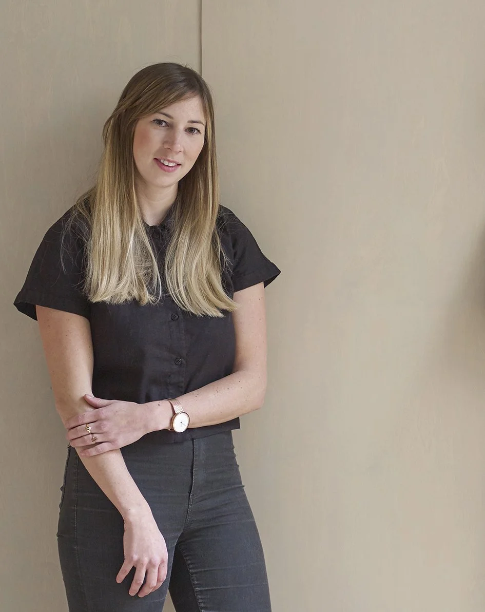 A young woman standing against a beige wall, wearing a black short-sleeve shirt and dark jeans, with long blonde hair and a watch on her left wrist.