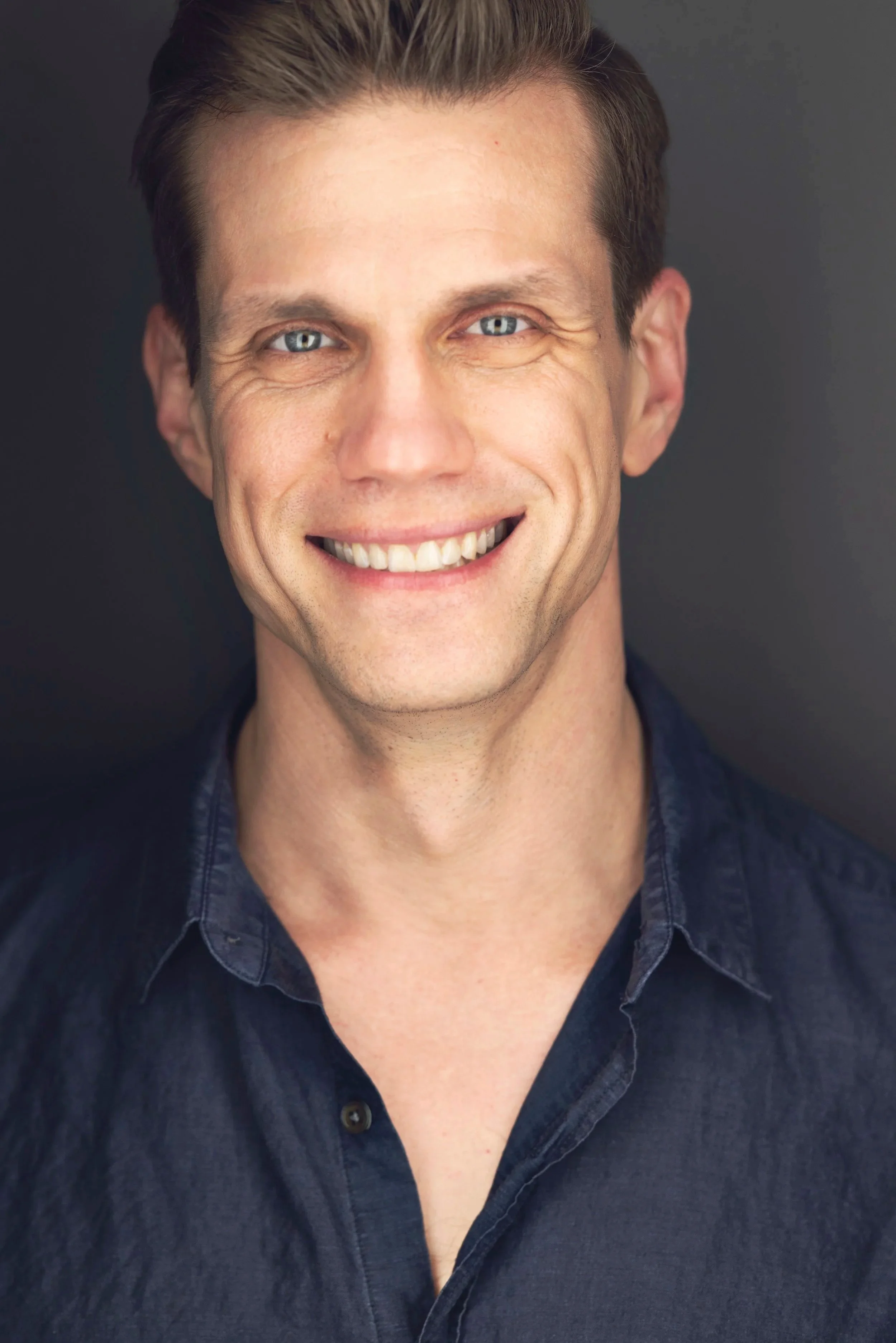 Close-up portrait of a smiling man with blue eyes and dark brown hair, wearing a dark blue button-up shirt against a plain dark background.