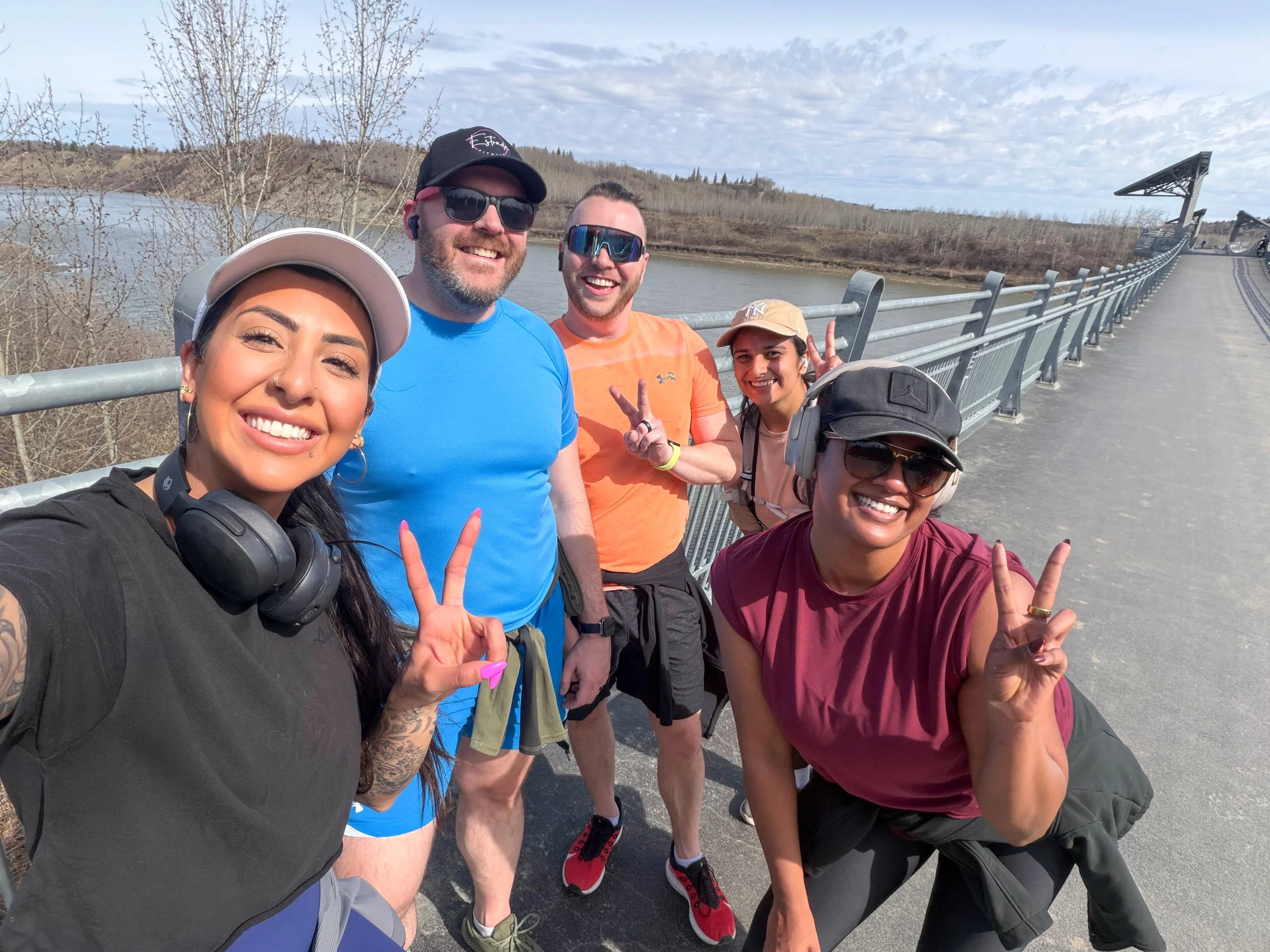 Group of five smiling people taking a selfie outdoors on a bridge over water with a partly cloudy sky.