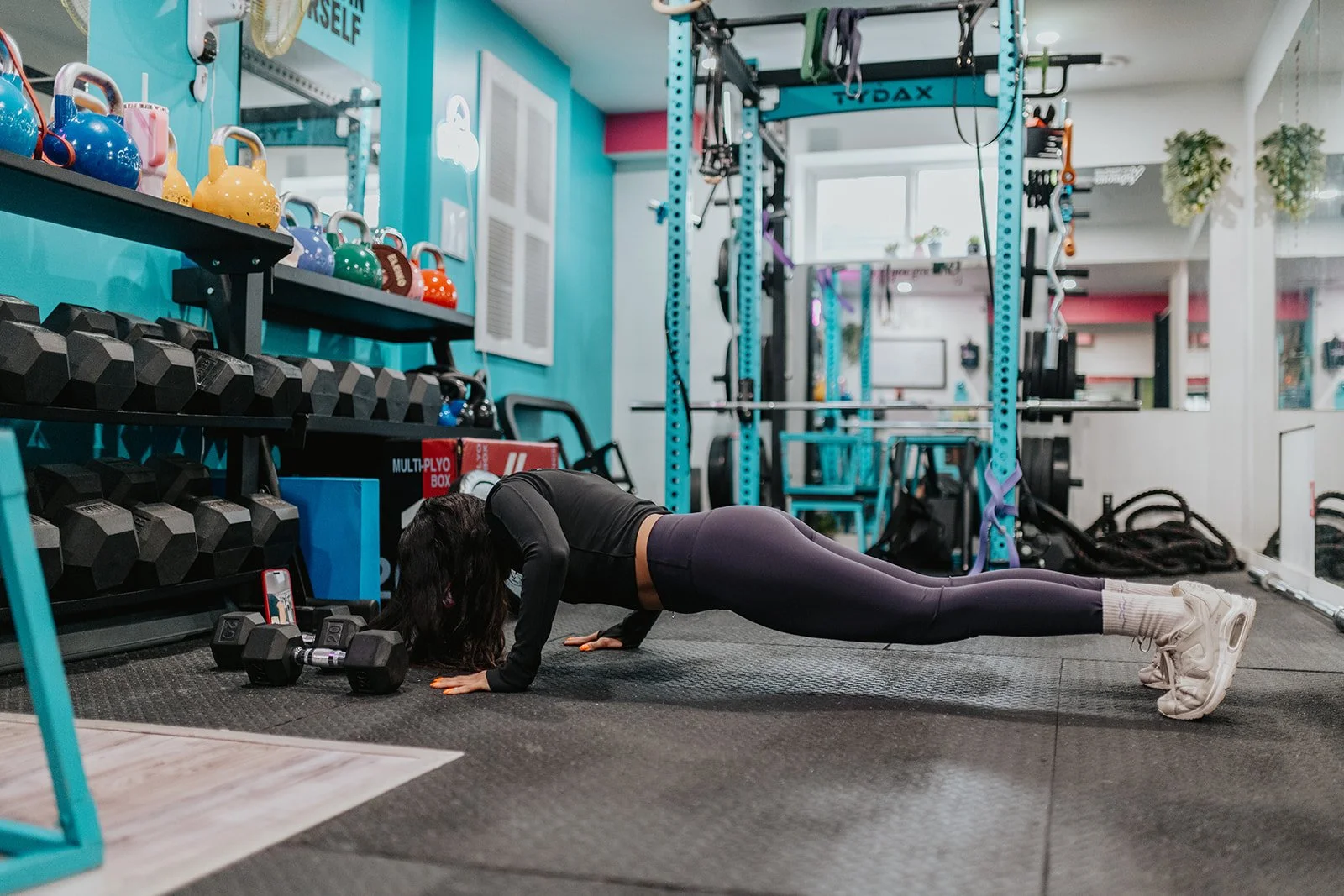 A woman in athletic wear doing a plank exercise in a gym, with weights and workout equipment visible around her.