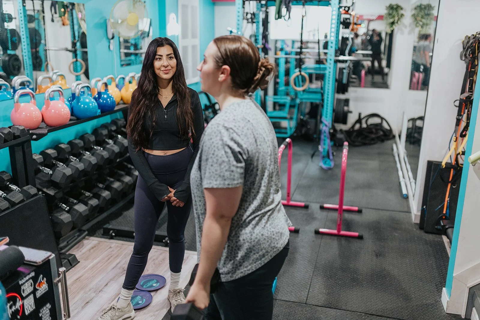 Two women talking in a gym with workout equipment, dumbbells, kettlebells, and exercise mats visible.